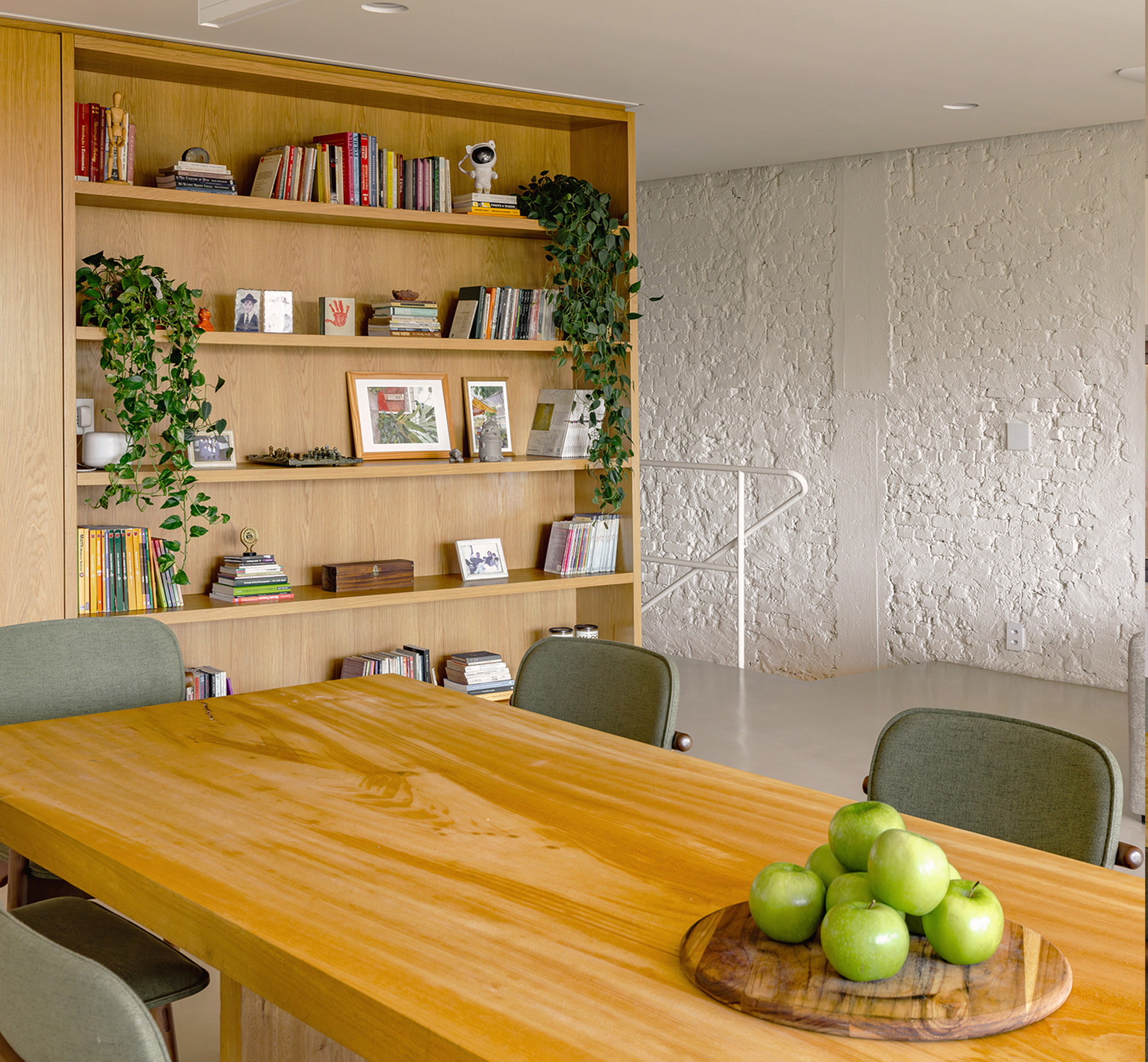 Oak shelving unit with books, framed prints and trailing plants beside a painted brick wall and white stair railing, green apples on the table