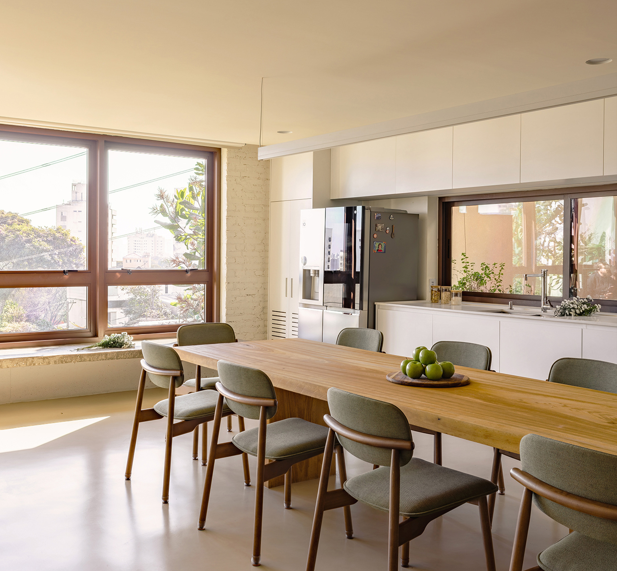 Long oak dining table with green upholstered chairs facing a white kitchen run and timber-framed windows in warm afternoon sun