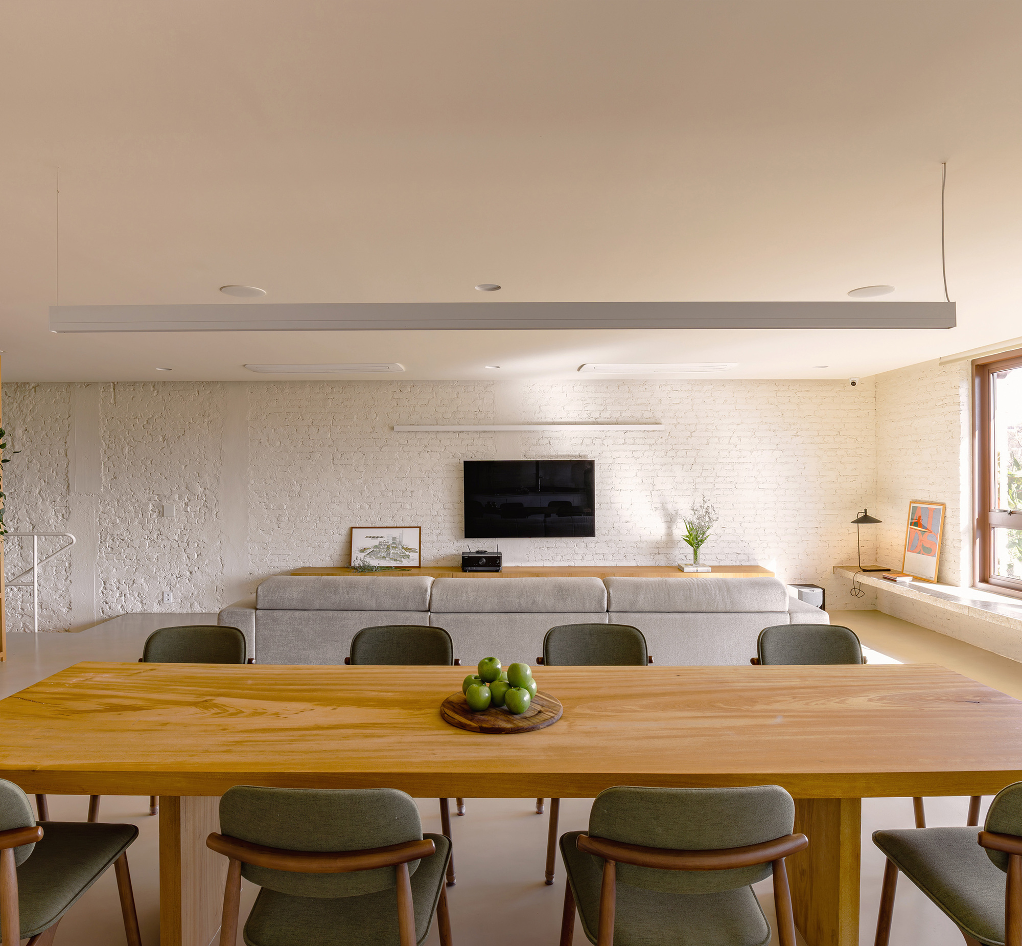 Dining table in the foreground with a grey sofa and wall-mounted television against a white painted brick wall under soft daylight