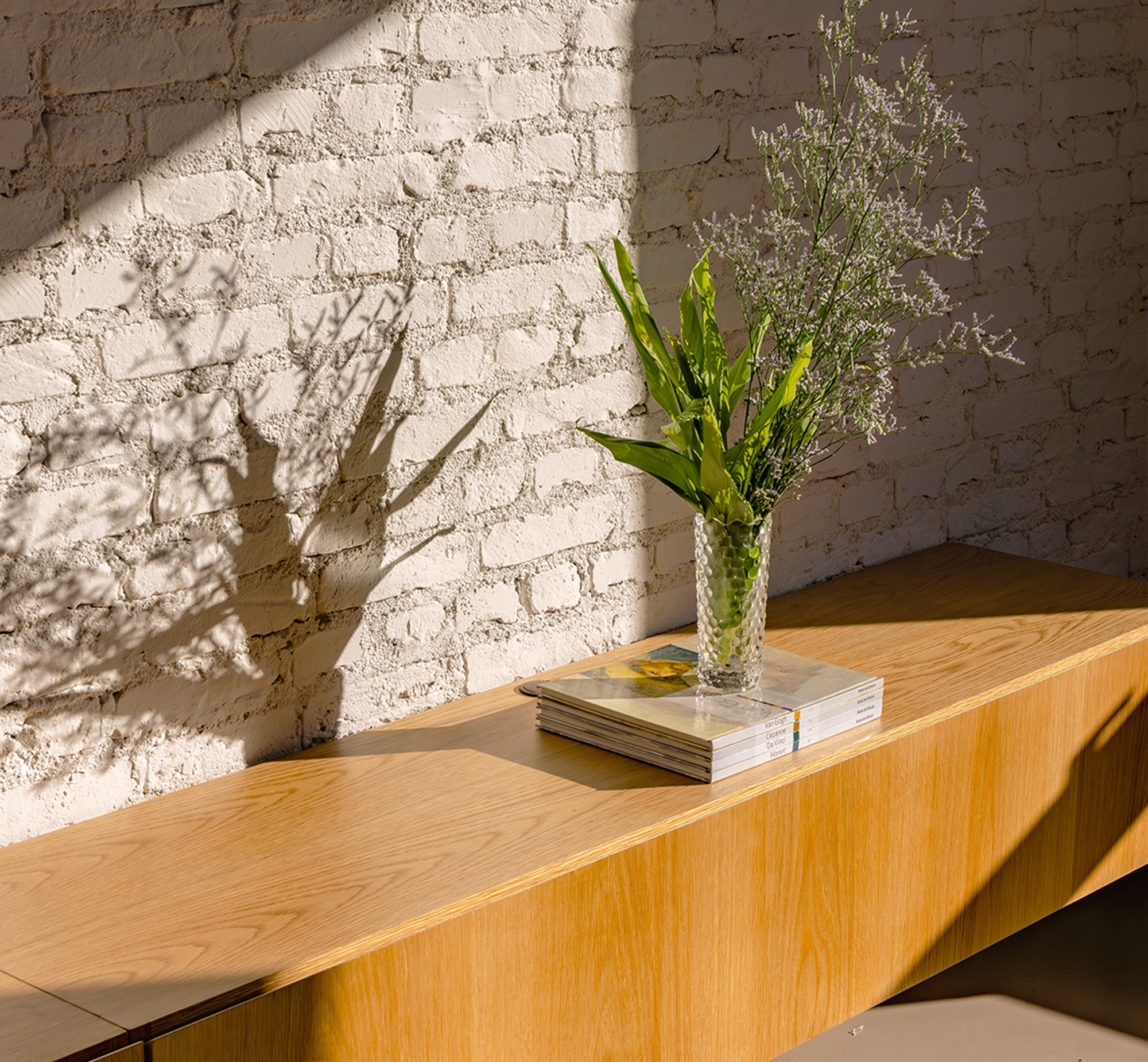 Oak ledge with stacked magazines and a vase of foliage against painted brick, tree shadows falling across the wall in low sun