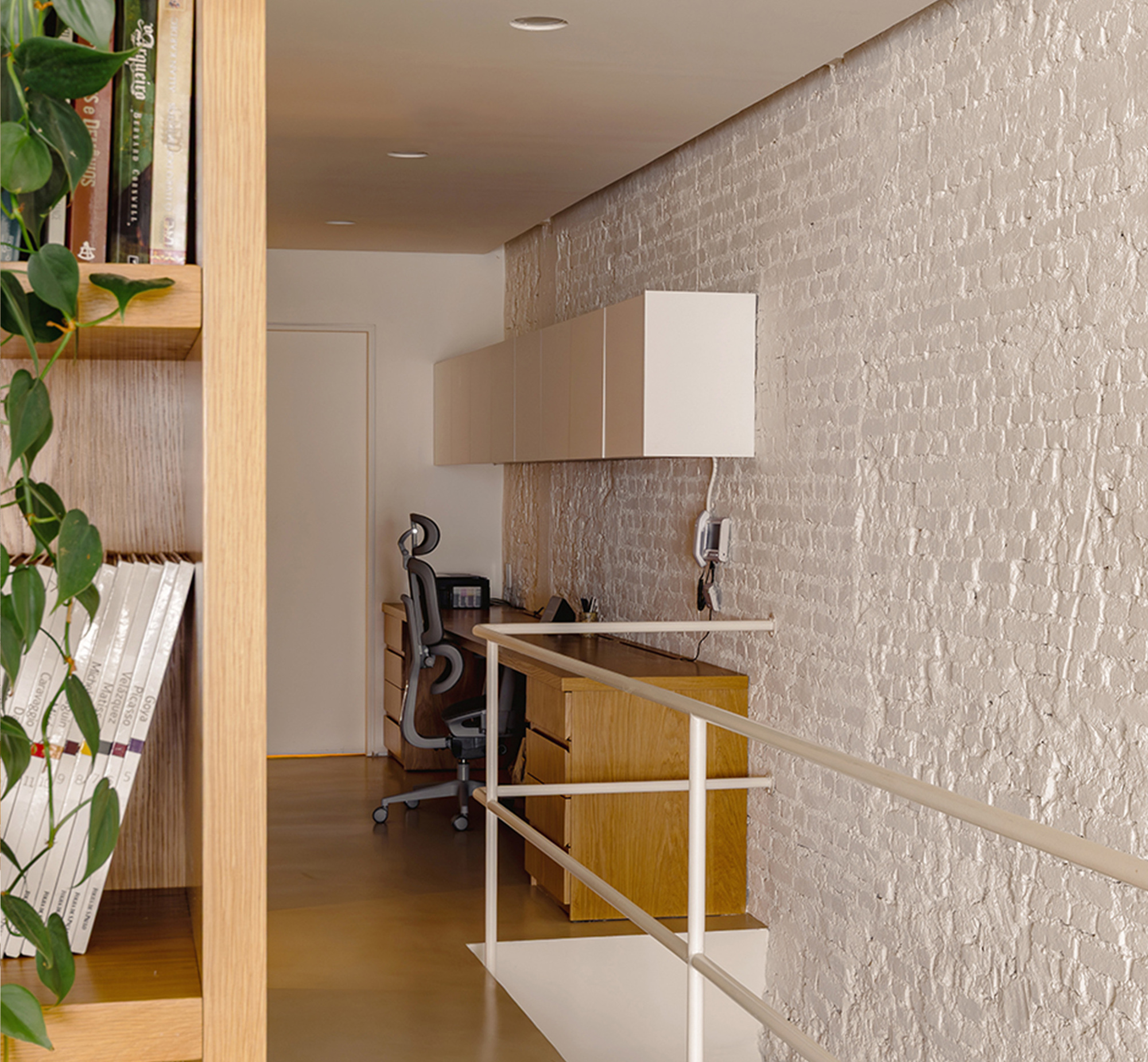 Narrow study nook with oak desk and wall cabinets behind a white metal railing, plant trailing from a shelf in the foreground