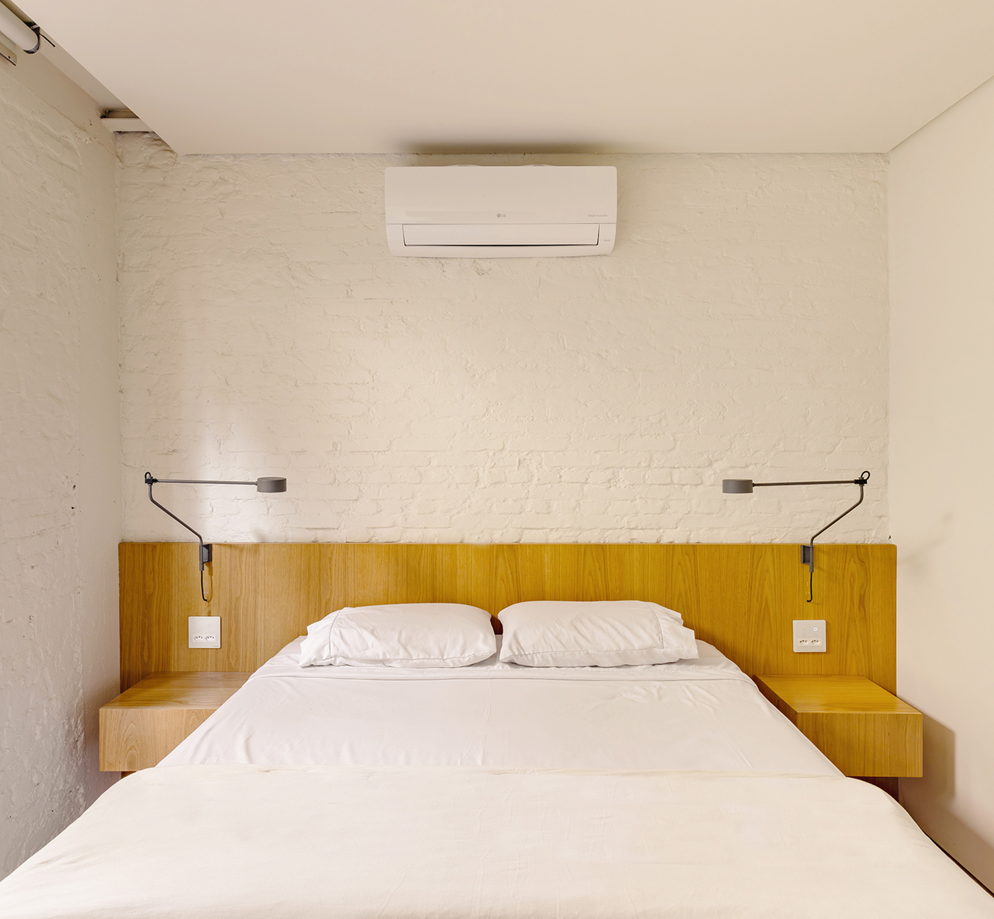 Bedroom with an oak headboard and floating bedside shelves against white painted brick, swing-arm lamps on either side of the bed