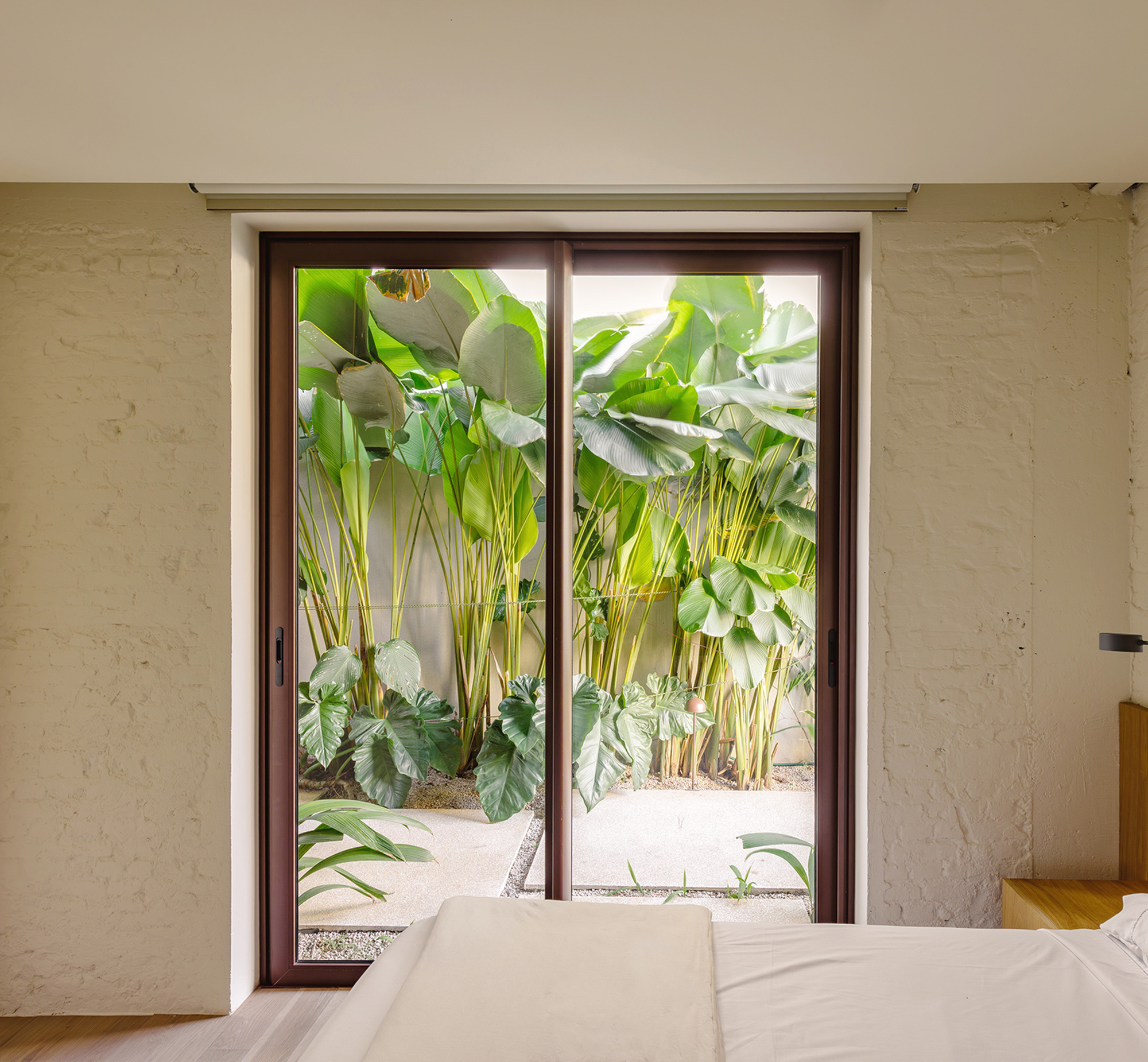 Bedroom sliding glass door framed in dark metal, opening onto a small courtyard of banana-leaf plants in soft daylight