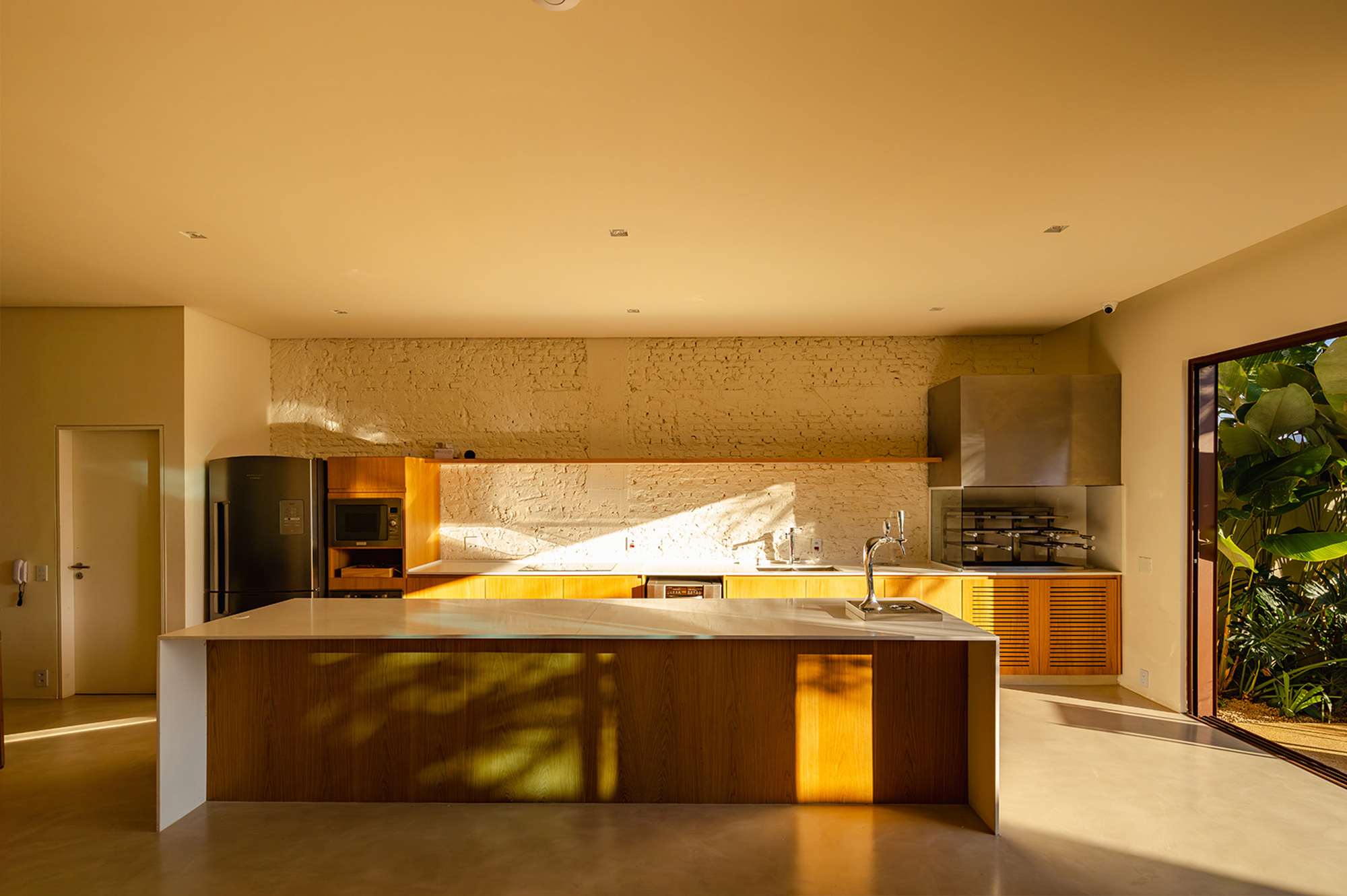 Kitchen with a long timber-clad island and whitewashed brick wall, patterned by low golden sunlight and leaf shadows