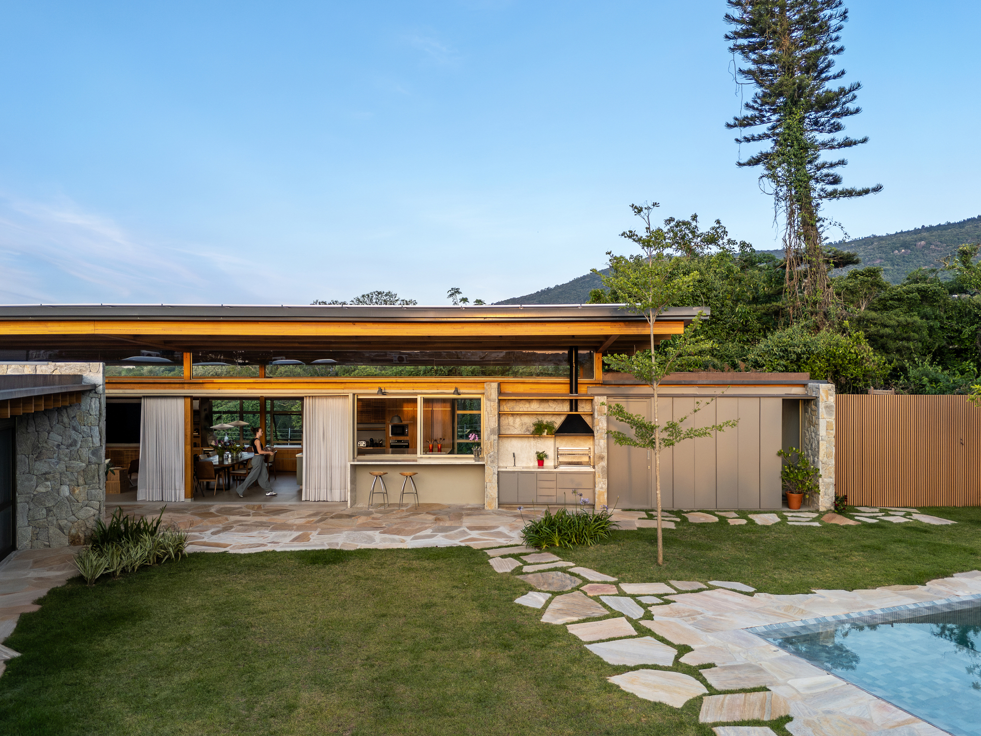 Low timber-edged roof over a glazed kitchen and dining pavilion, with a stone-paved lawn path leading to the pool at dusk