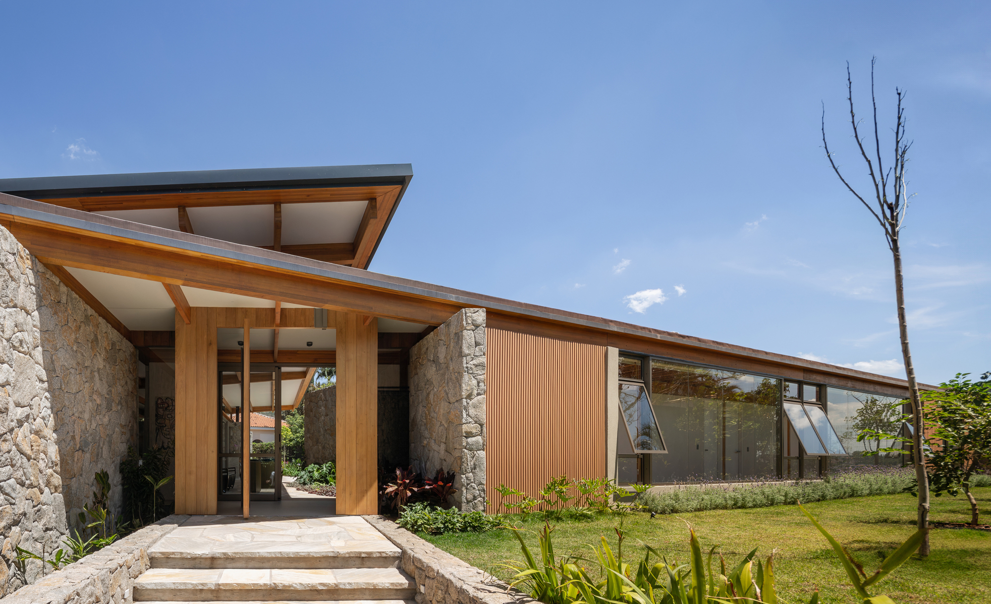 Entrance porch with timber columns and a stone wall, beside a slatted wood panel and glazed band under a blue sky