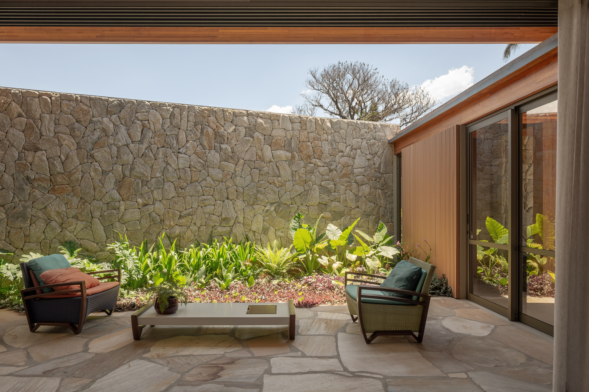 Paved patio with two lounge chairs and a low table facing a rubble stone wall and a planted bed of tropical foliage