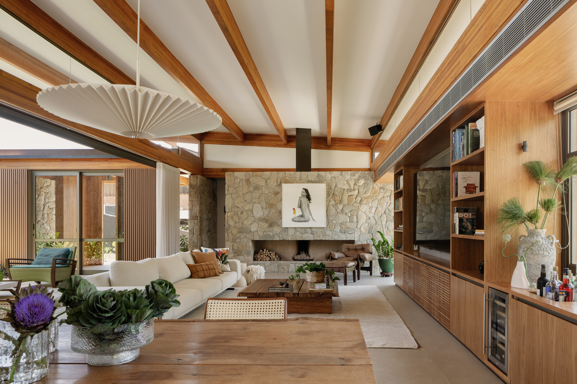 Living room with exposed timber beams, a stone fireplace wall and built-in wood shelving, with a pleated pendant lamp in the foreground