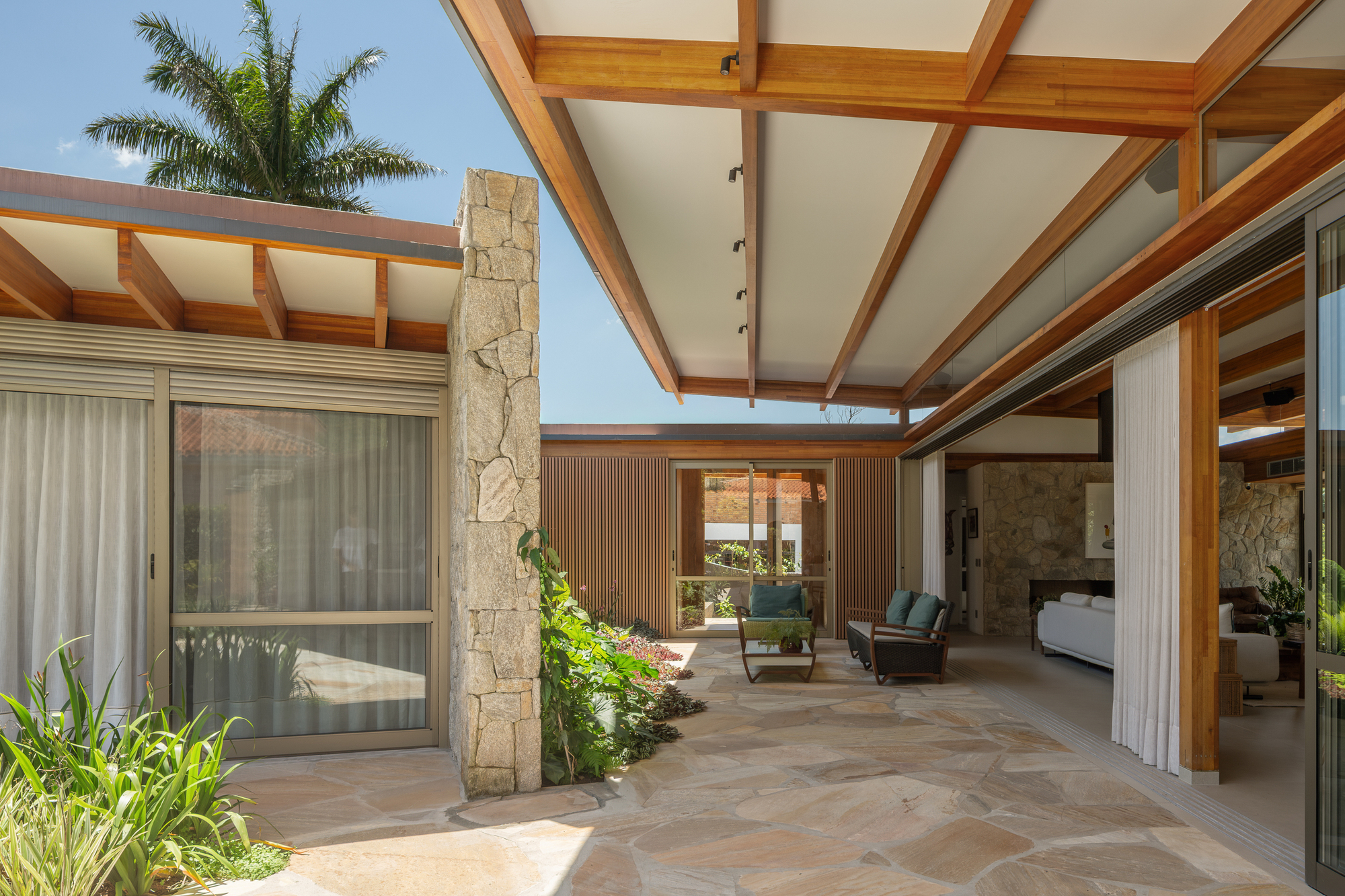 Covered courtyard of irregular flagstone paving, a stone pier and timber roof beams, with a palm rising behind in bright daylight