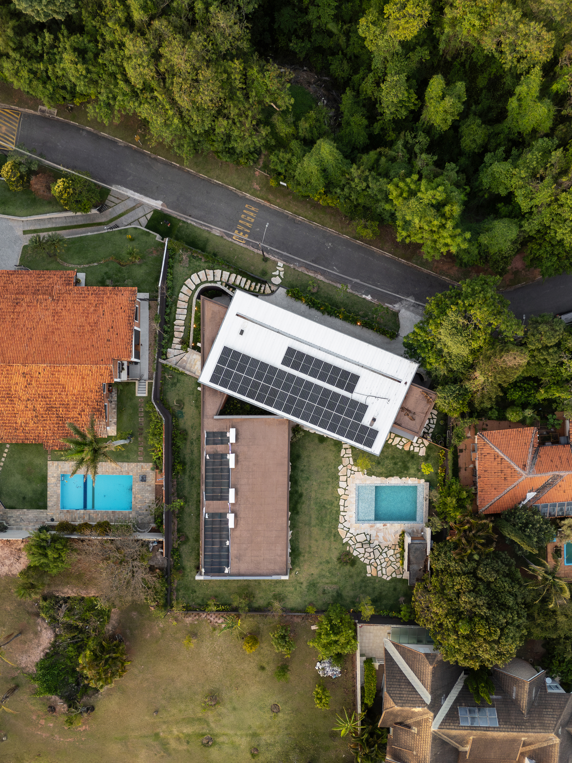 Drone view of the white roof with solar panels and a second brown roof, flanked by a pool, road and forest