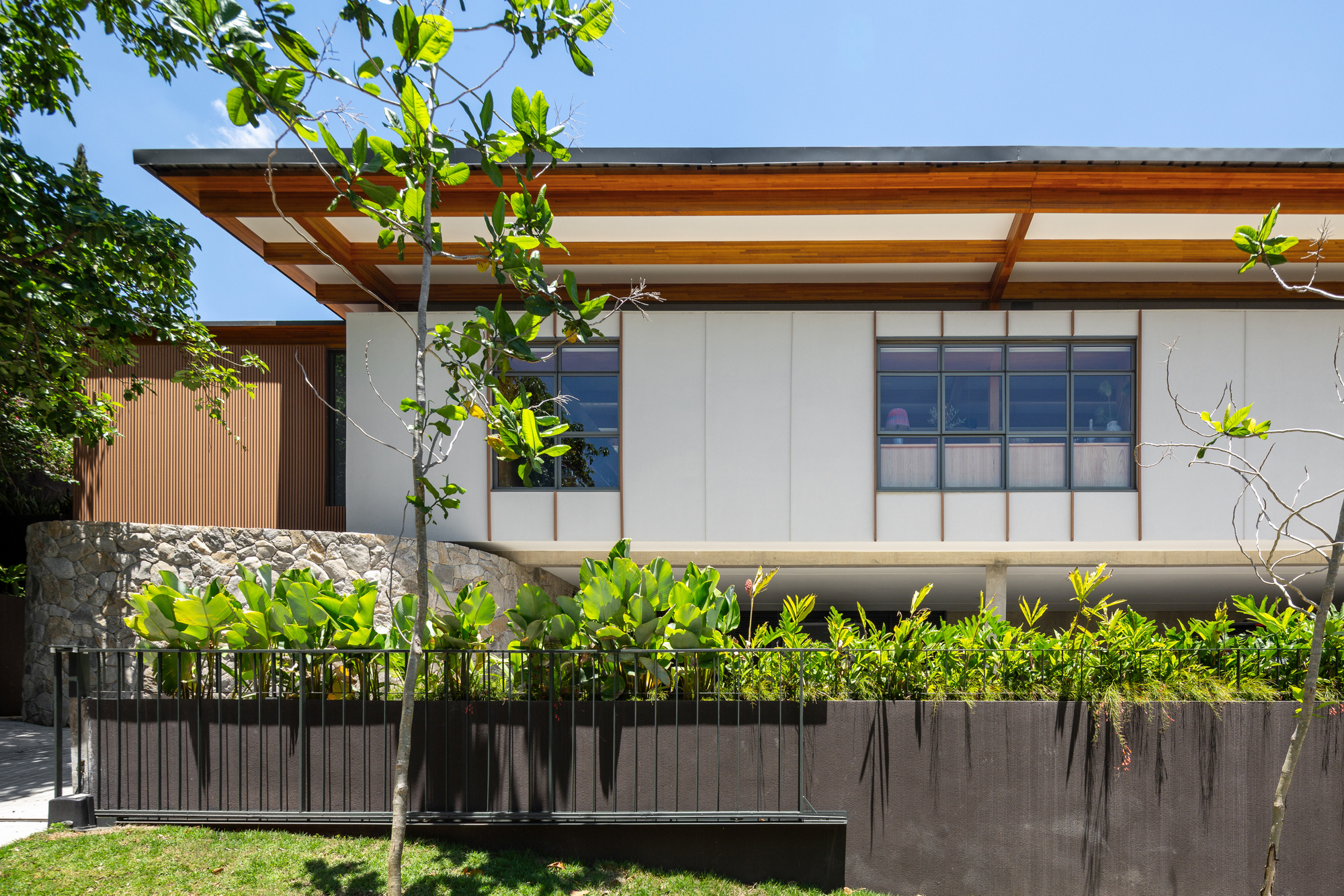 Street-side facade with a deep timber-eaved roof over white panels, a stone wall and a planted dark concrete bed in bright sun