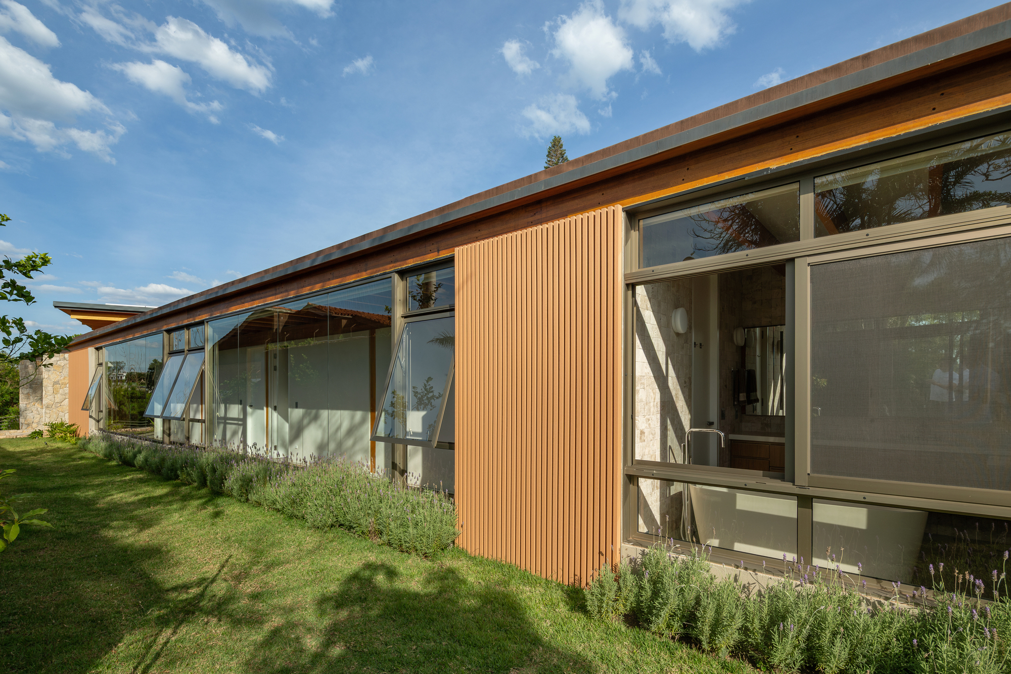 Side elevation with a slatted timber panel between awning windows, lavender along the lawn edge under a blue sky with scattered clouds