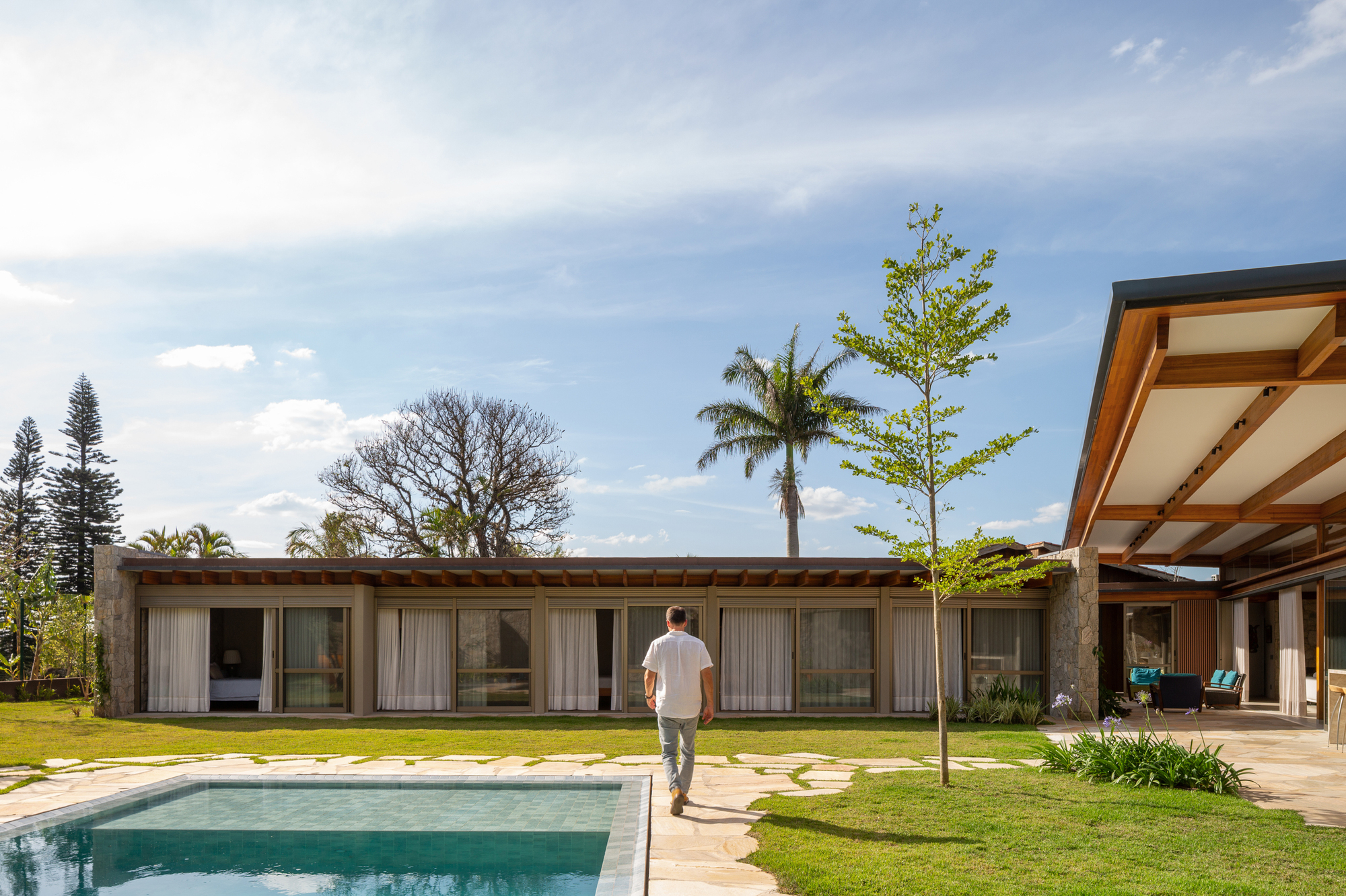 Single-storey wing with curtained glazing beyond a pool, a man in a white shirt walking across the stone paving toward it