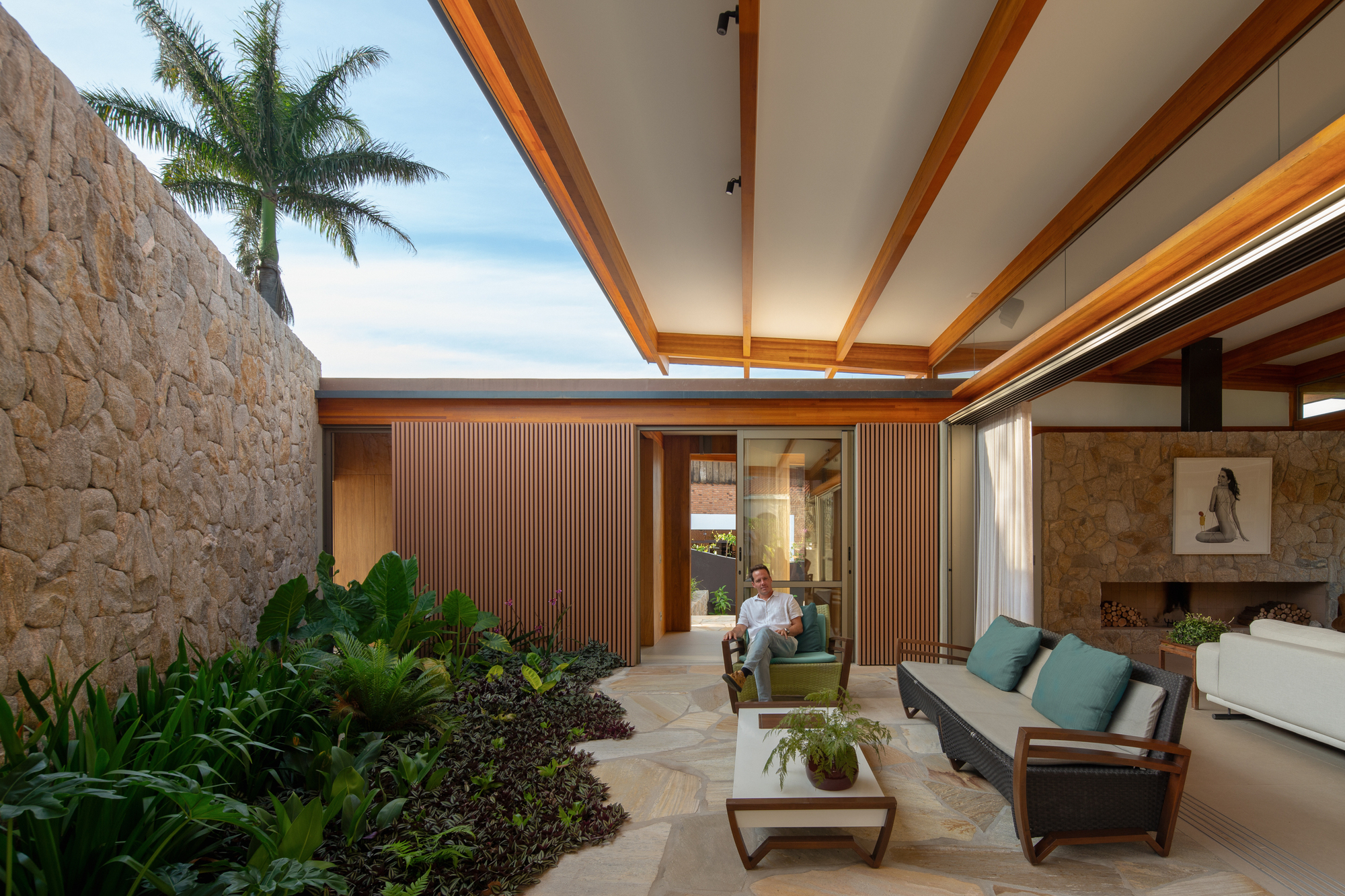 Covered terrace with exposed timber beams, a rough stone wall and a planted bed, a seated man beside the slatted doors