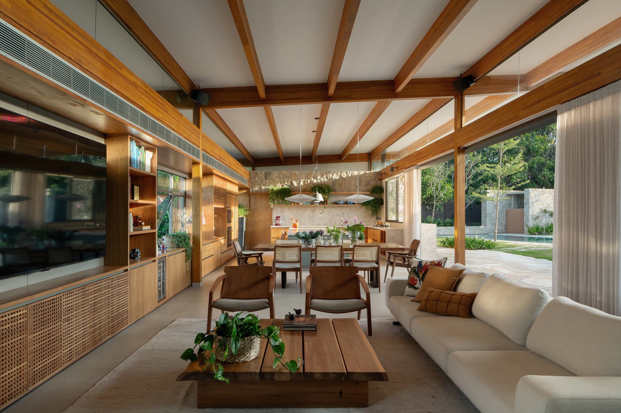 Living and dining space under exposed timber beams, with built-in joinery on the left and a garden and pool glimpsed through sheer curtains
