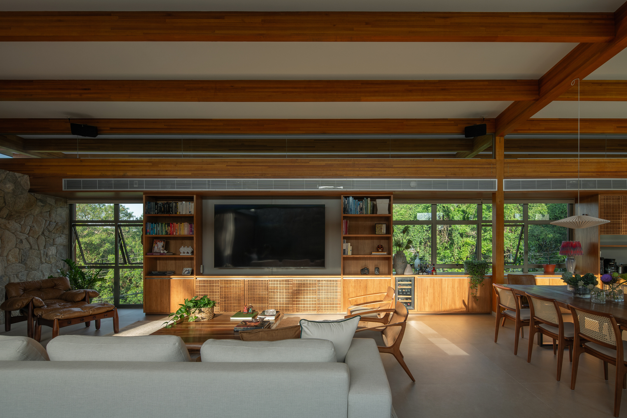 Living room view toward a timber media wall with shelving, framed by glazing that looks onto dense green trees in afternoon light