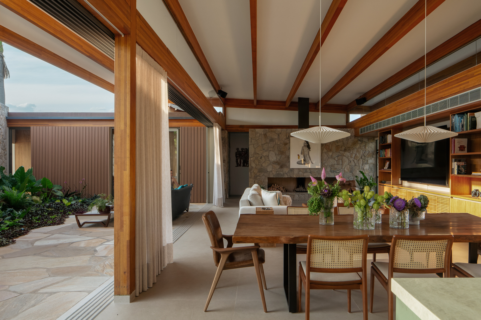 Open living and dining area with timber beams, stone fireplace wall and sheer curtain facing a planted stone-paved courtyard at dusk