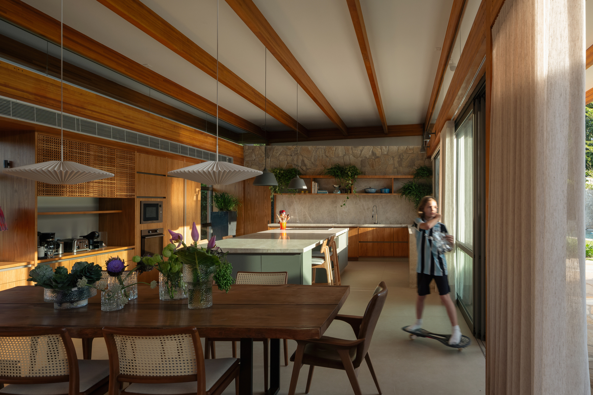 Dining table with flower vases in the foreground and a child on a skateboard near a sheer curtain, kitchen beyond