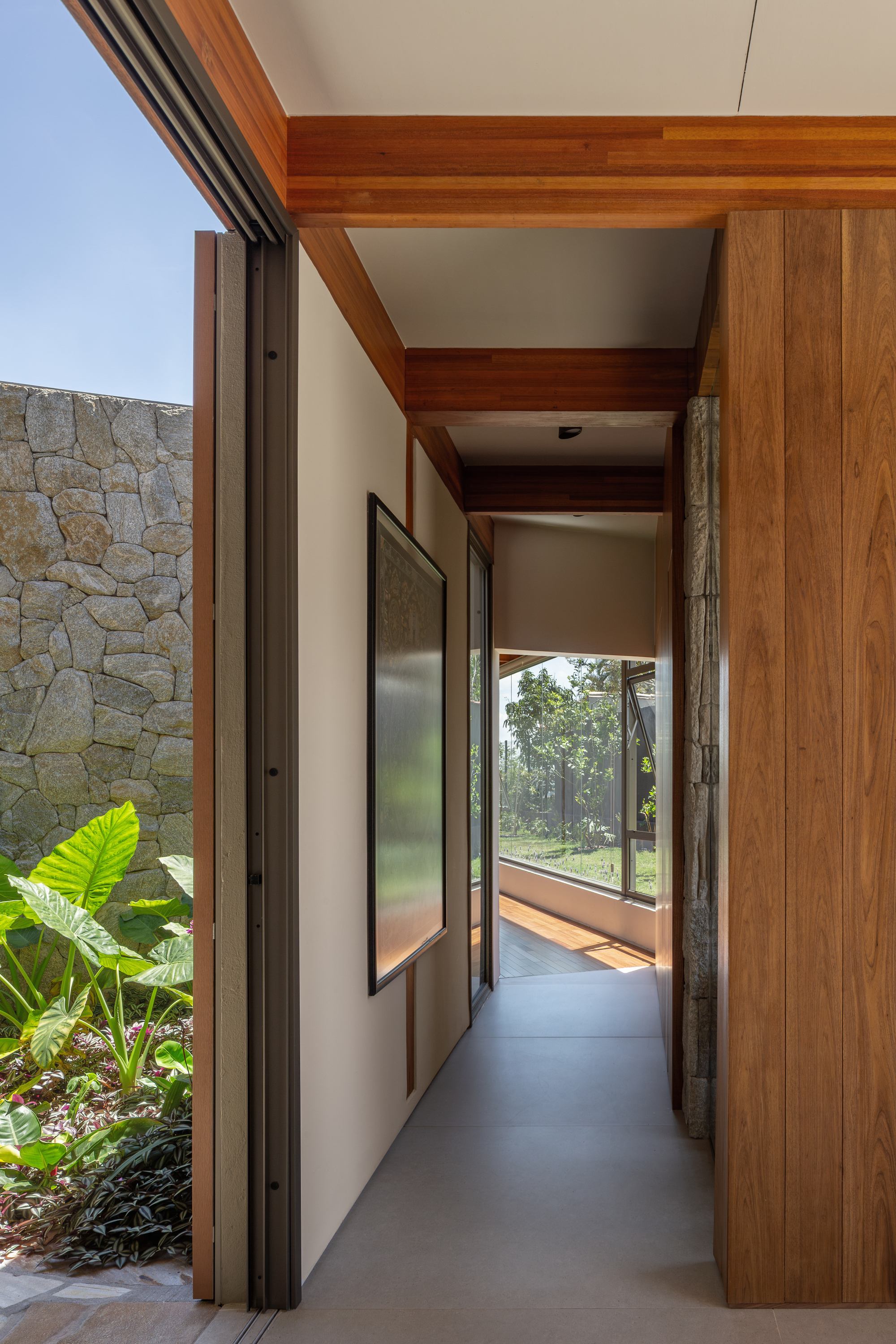 Narrow corridor with timber beams and wood-paneled wall, opening on the left to a stone wall and large-leaf plants in sunlight