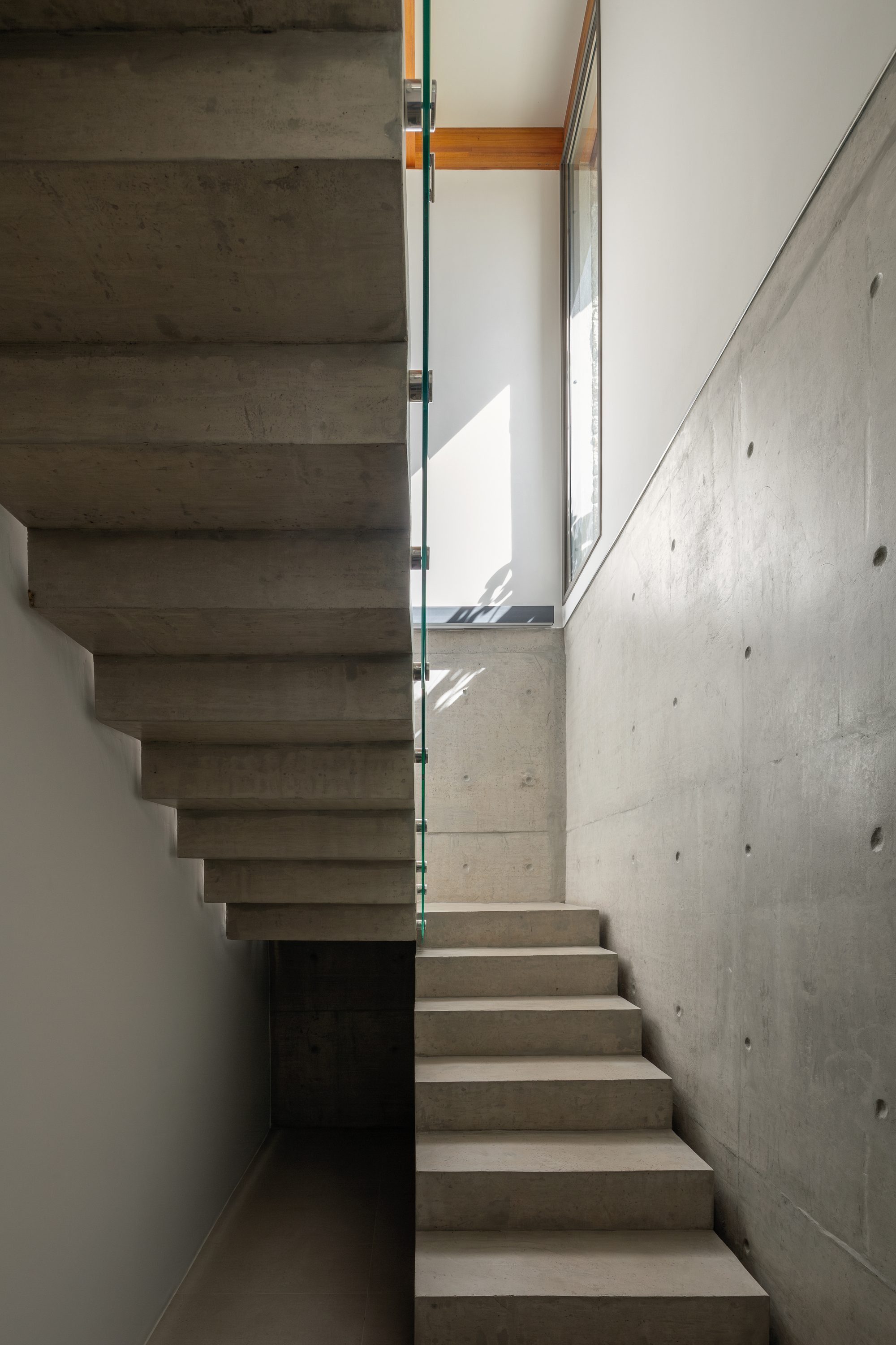 Cantilevered concrete stair with board-marked walls and a glass balustrade, lit by a timber-framed window with sunlight on the wall