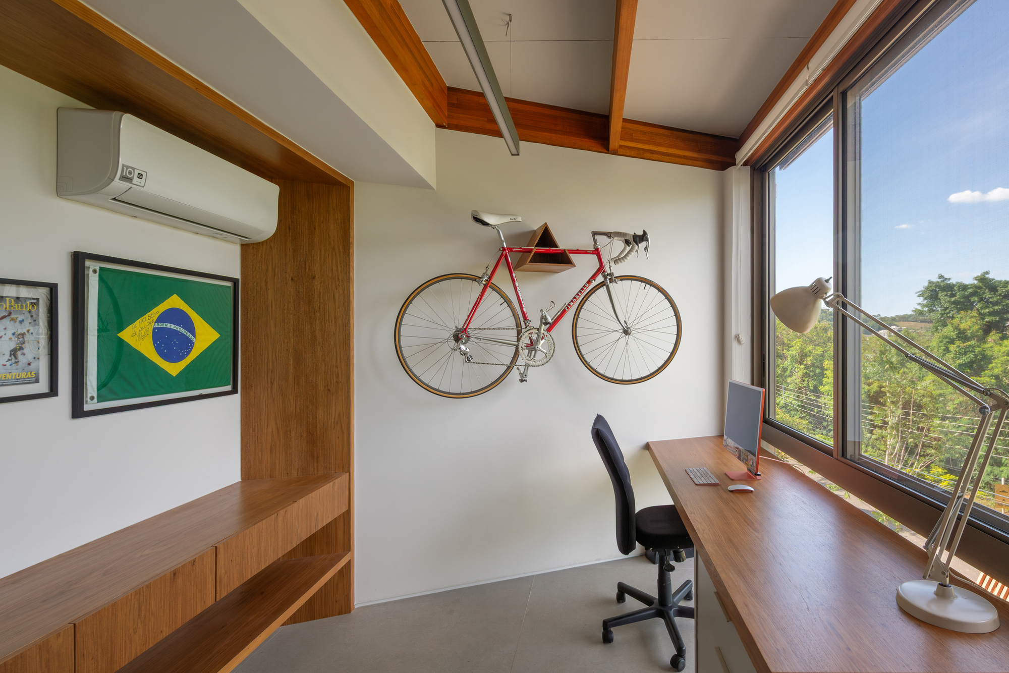 Home office with a red road bicycle mounted on the wall above a long timber desk, green hillside visible outside