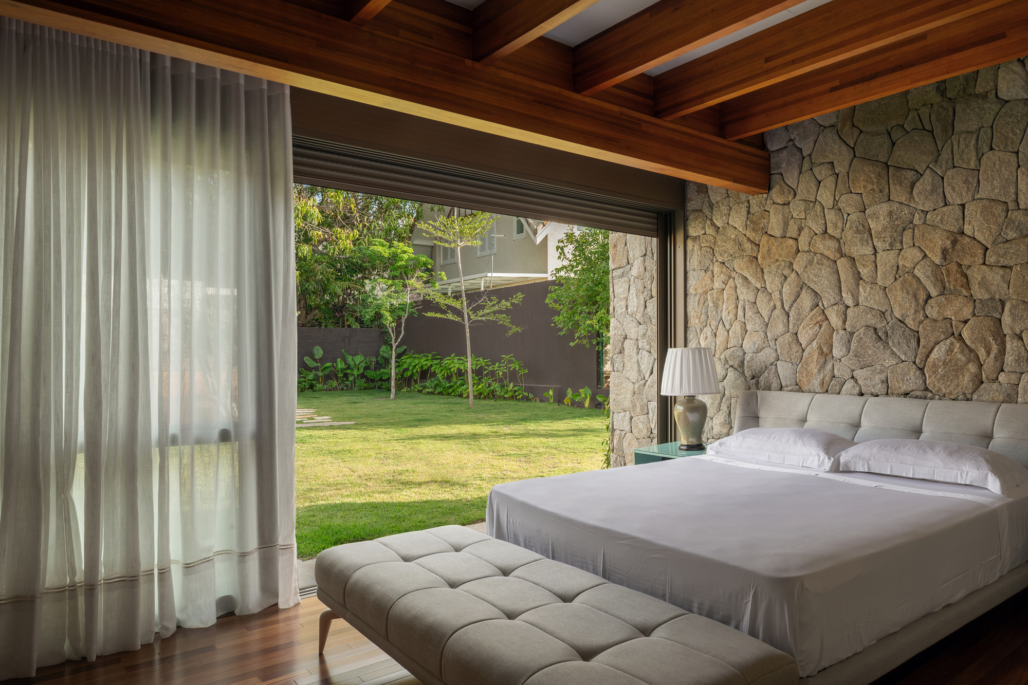 Bedroom with a rough stone wall, exposed timber beams and sheer curtains opening onto a sunlit lawn