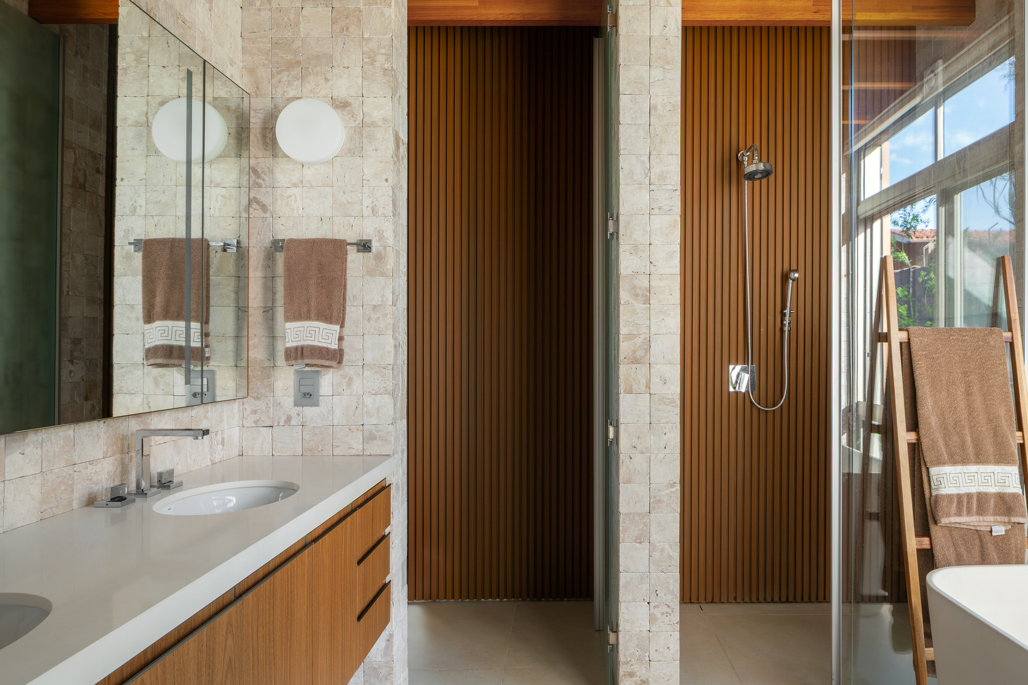 Bathroom with a timber vanity, travertine tile walls and a slatted wood shower wall, daylight coming through the glass screen