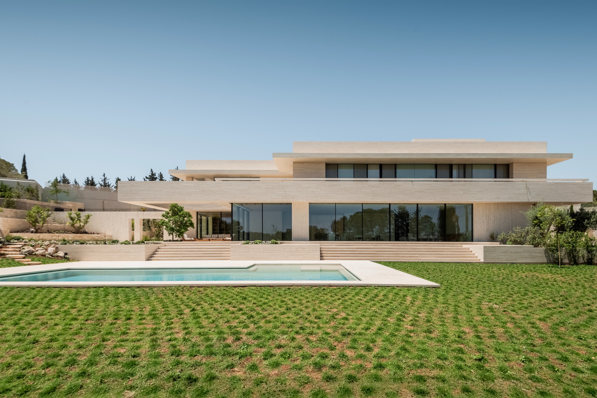 Stacked travertine volumes with deep cantilevered floors above a rectangular pool and a lawn of grass pavers under a clear sky
