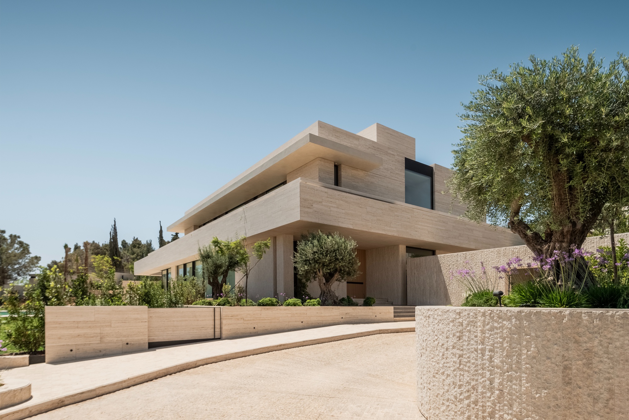 Layered stone facade seen from a curving drive, with an olive tree and purple agapanthus beside a rough-cut stone planter
