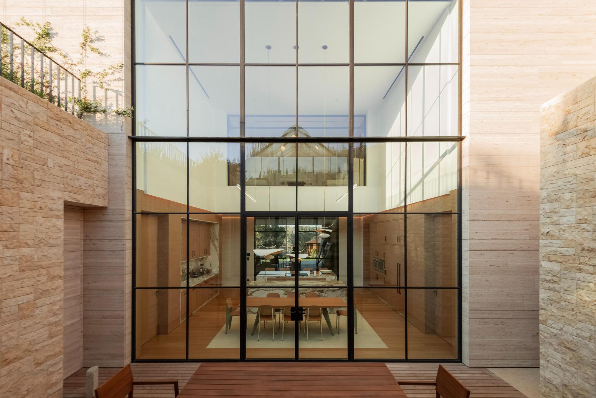 Double-height steel-framed glass wall opening to a dining room and kitchen, viewed across a timber deck between stone walls