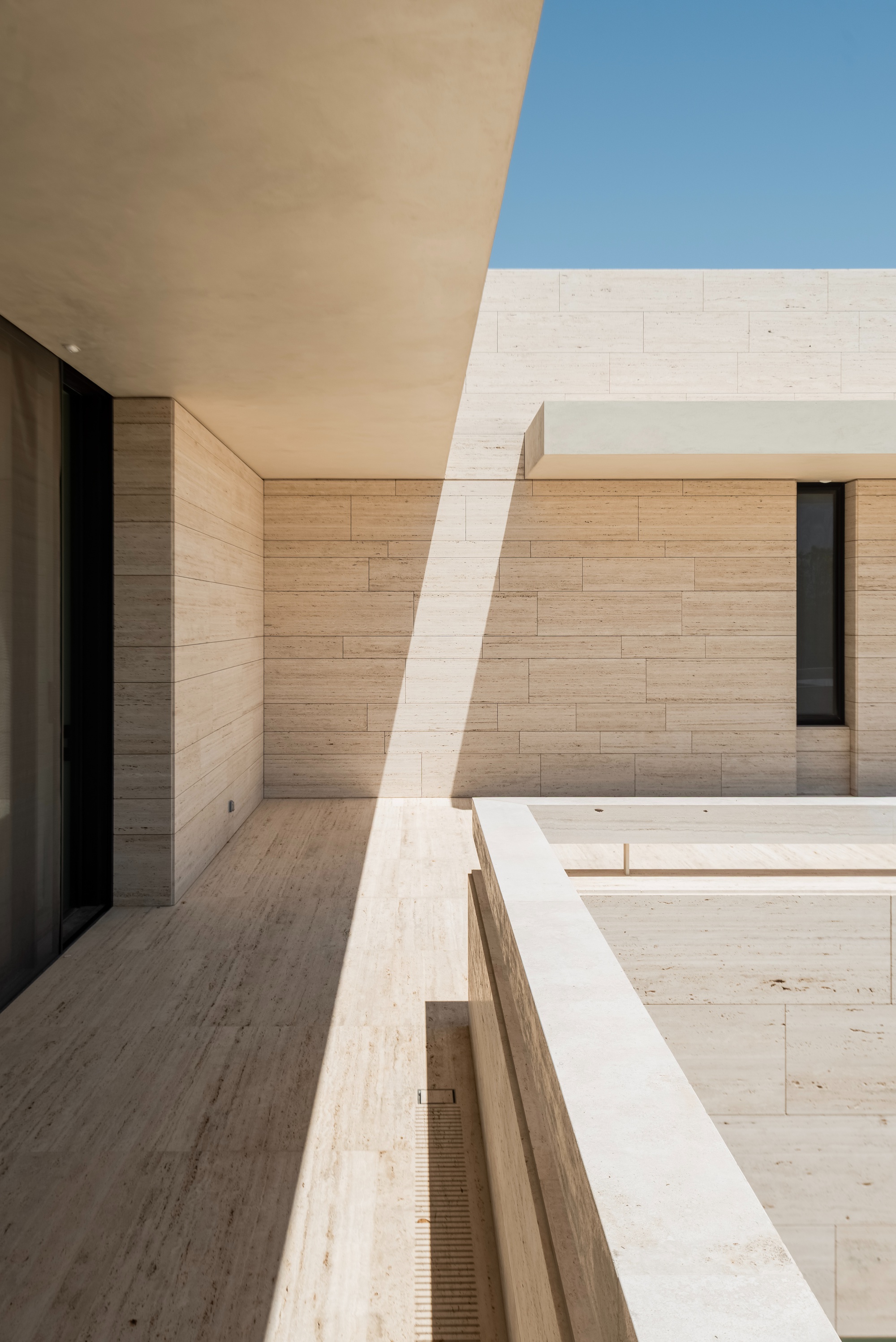 Covered terrace with a stone-clad wall, a slim vertical window and a pool edge, hard sunlight casting a diagonal shadow
