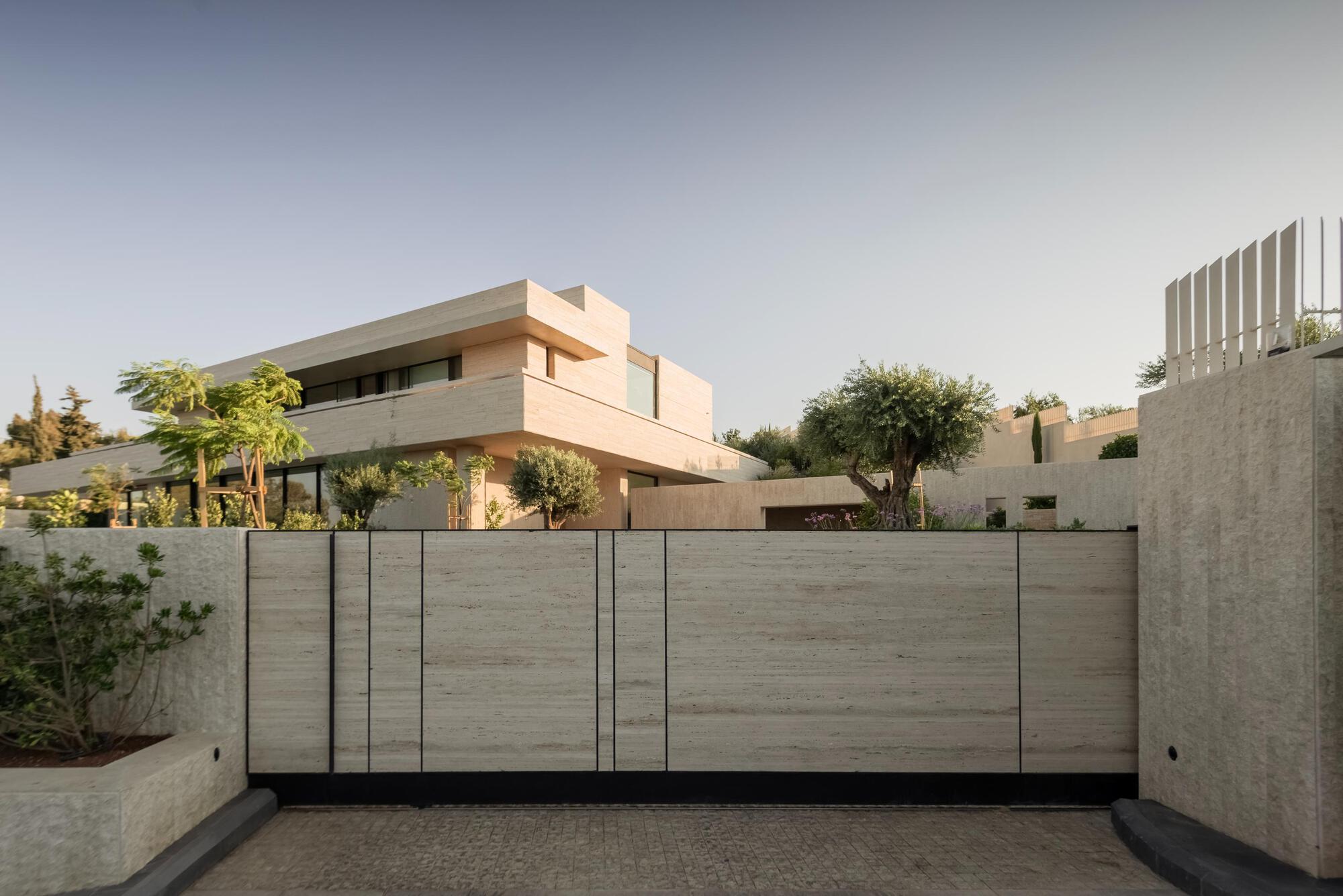 Street-side travertine gate and boundary wall with an olive tree and stacked upper volumes behind, under a clear dusk sky