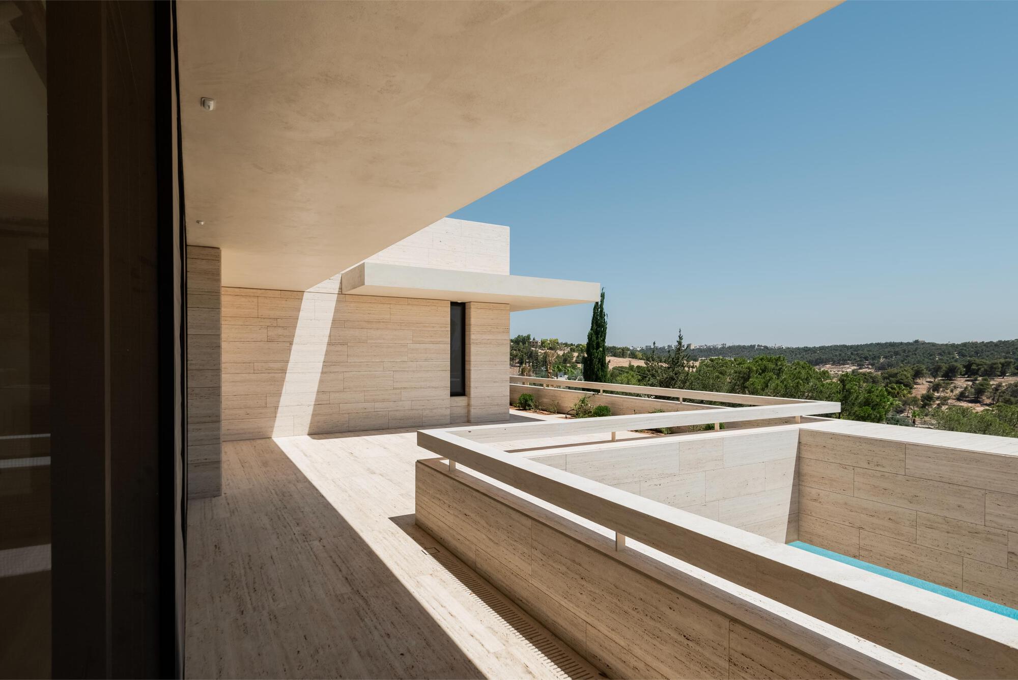 Upper terrace with travertine floor and low parapet, a sharp diagonal shadow cast across the wall and a pool edge beyond