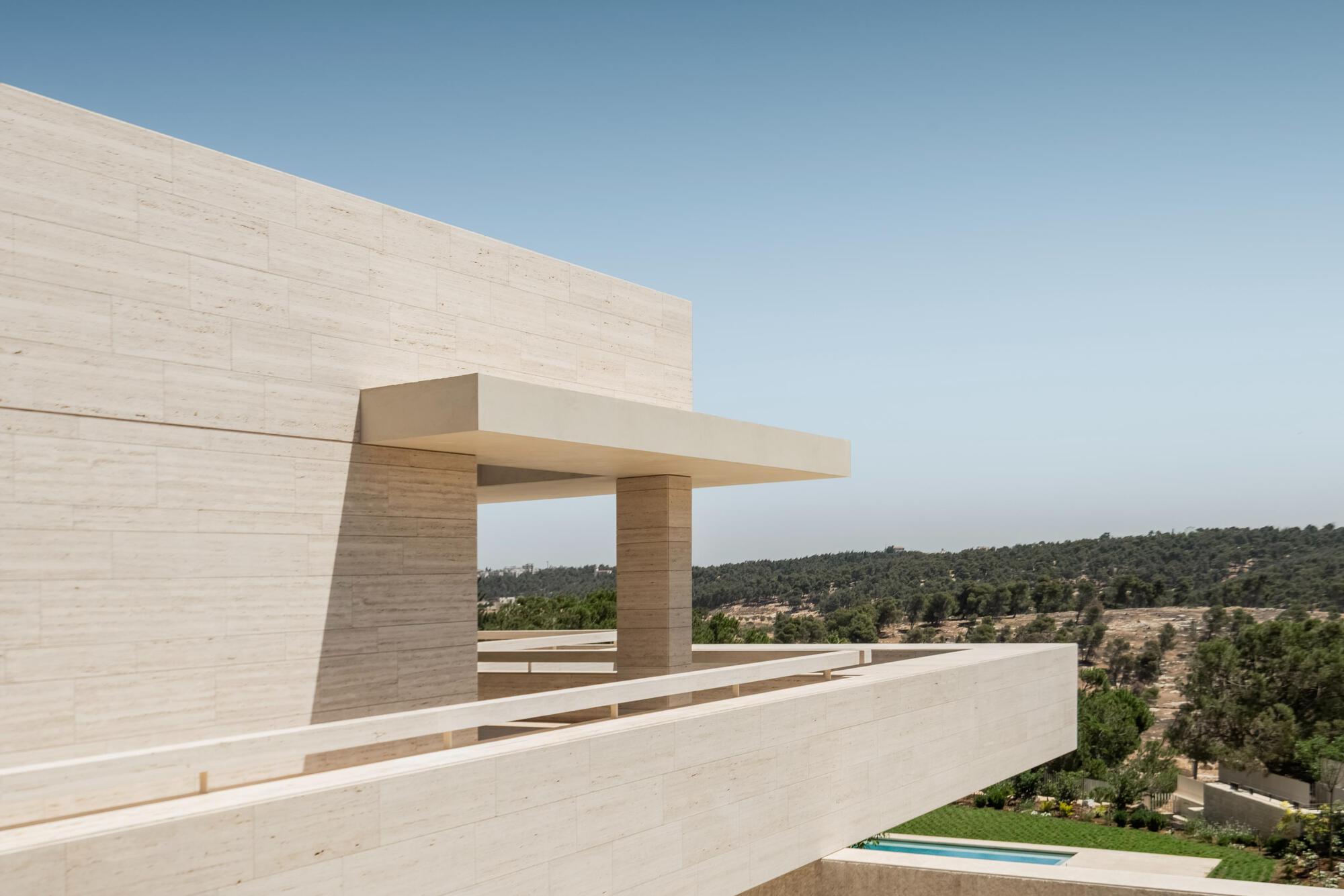 Cantilevered travertine terrace edge with a slim canopy and square pier, overlooking pine woodland and a distant pool