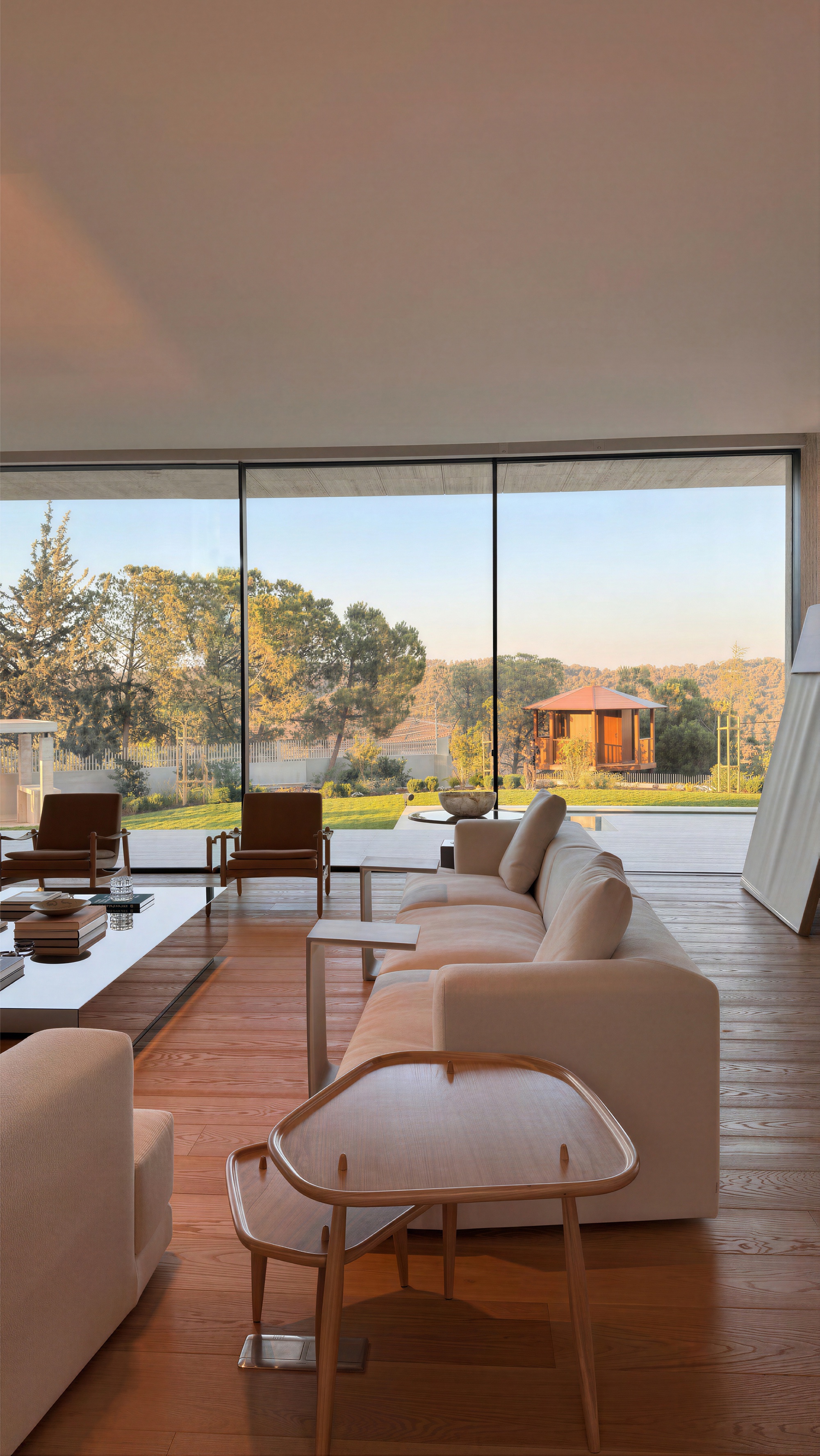 Living room with a cream sofa and timber floor facing full-height glazing and a garden pavilion in low evening light