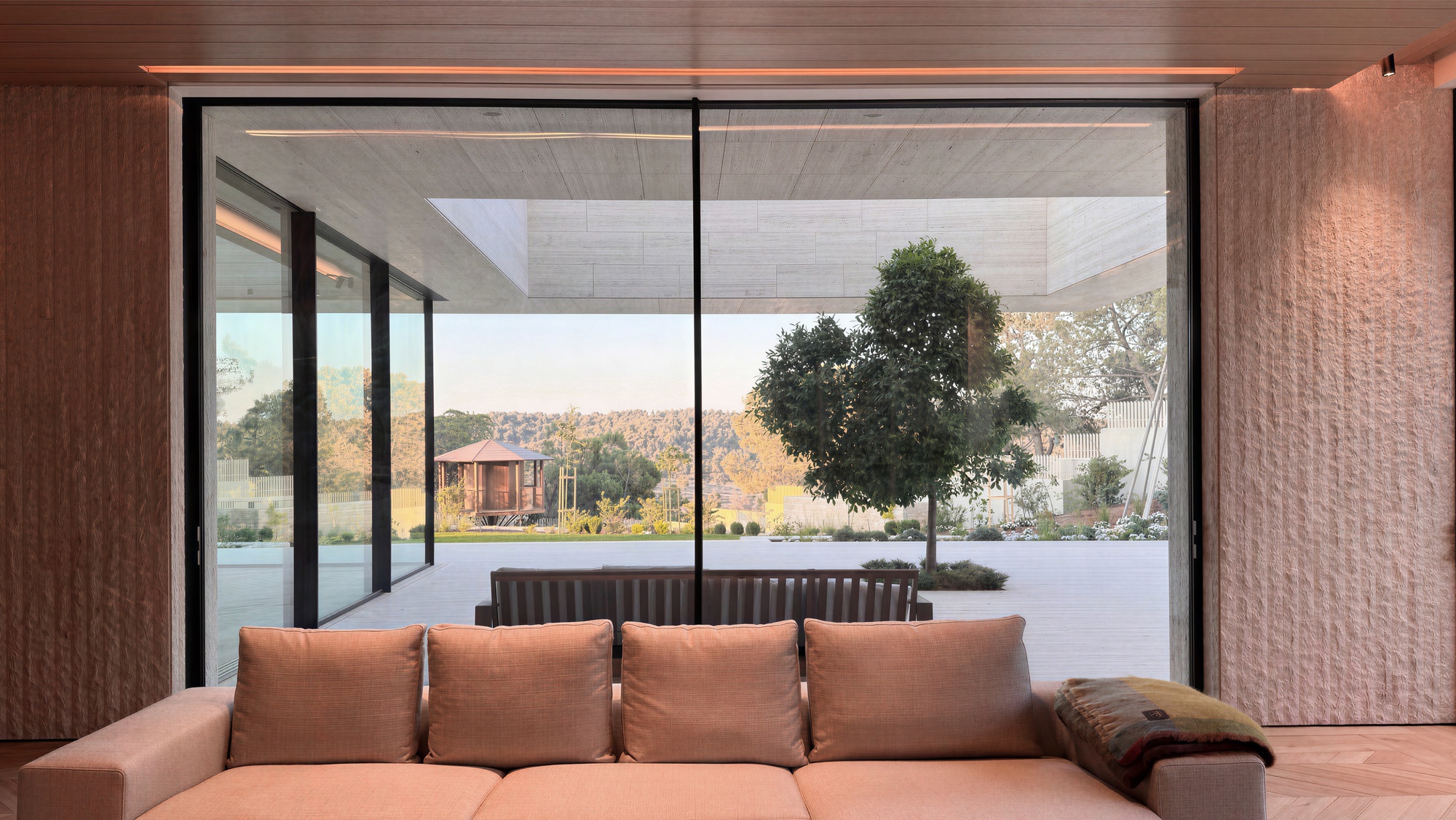 Floor-to-ceiling glass wall framing a lone tree, terrace and timber pavilion beyond a peach sofa in late afternoon light