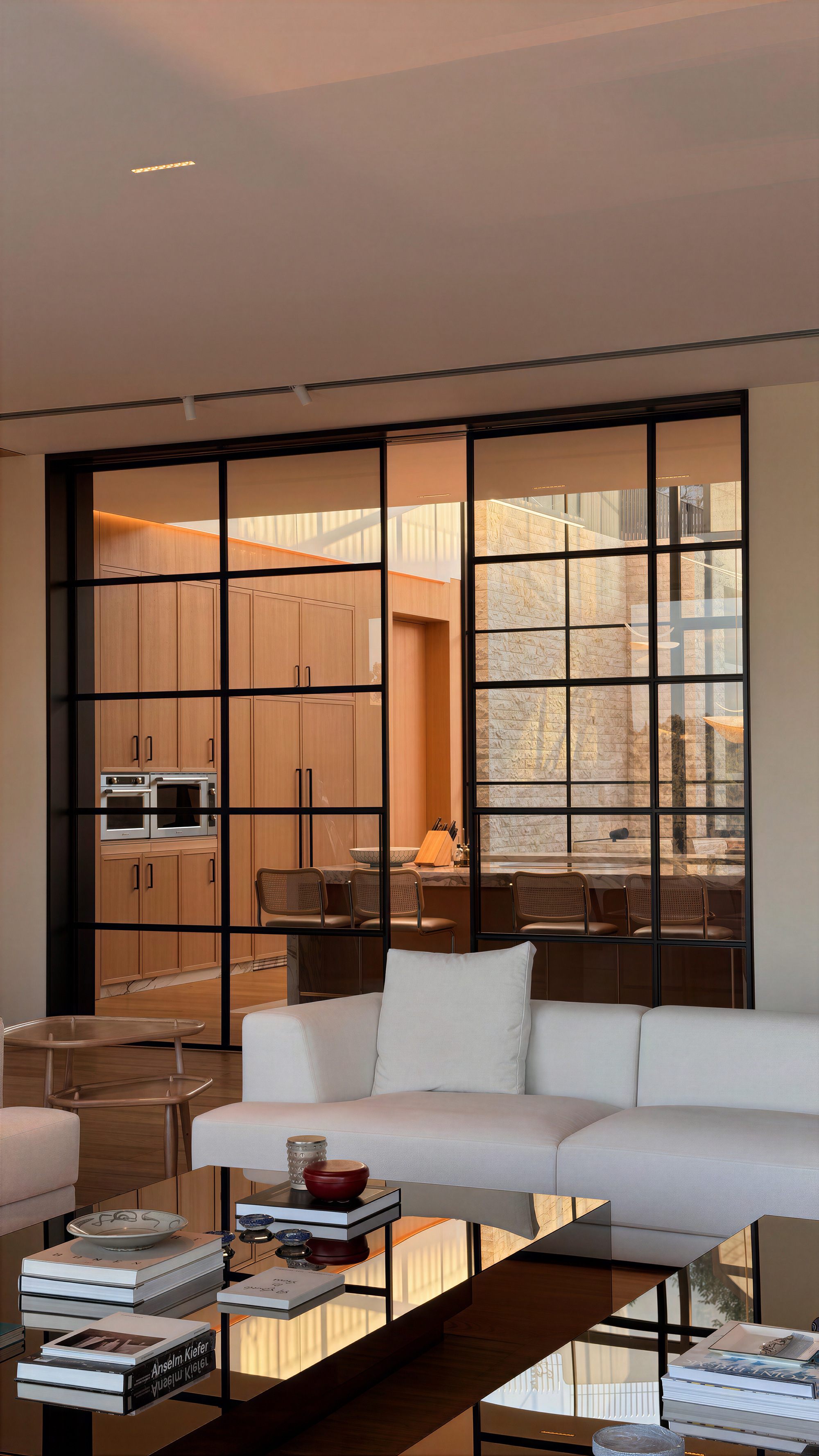 Black steel-framed glass partition between a white sofa and a timber kitchen, with books stacked on a glossy table