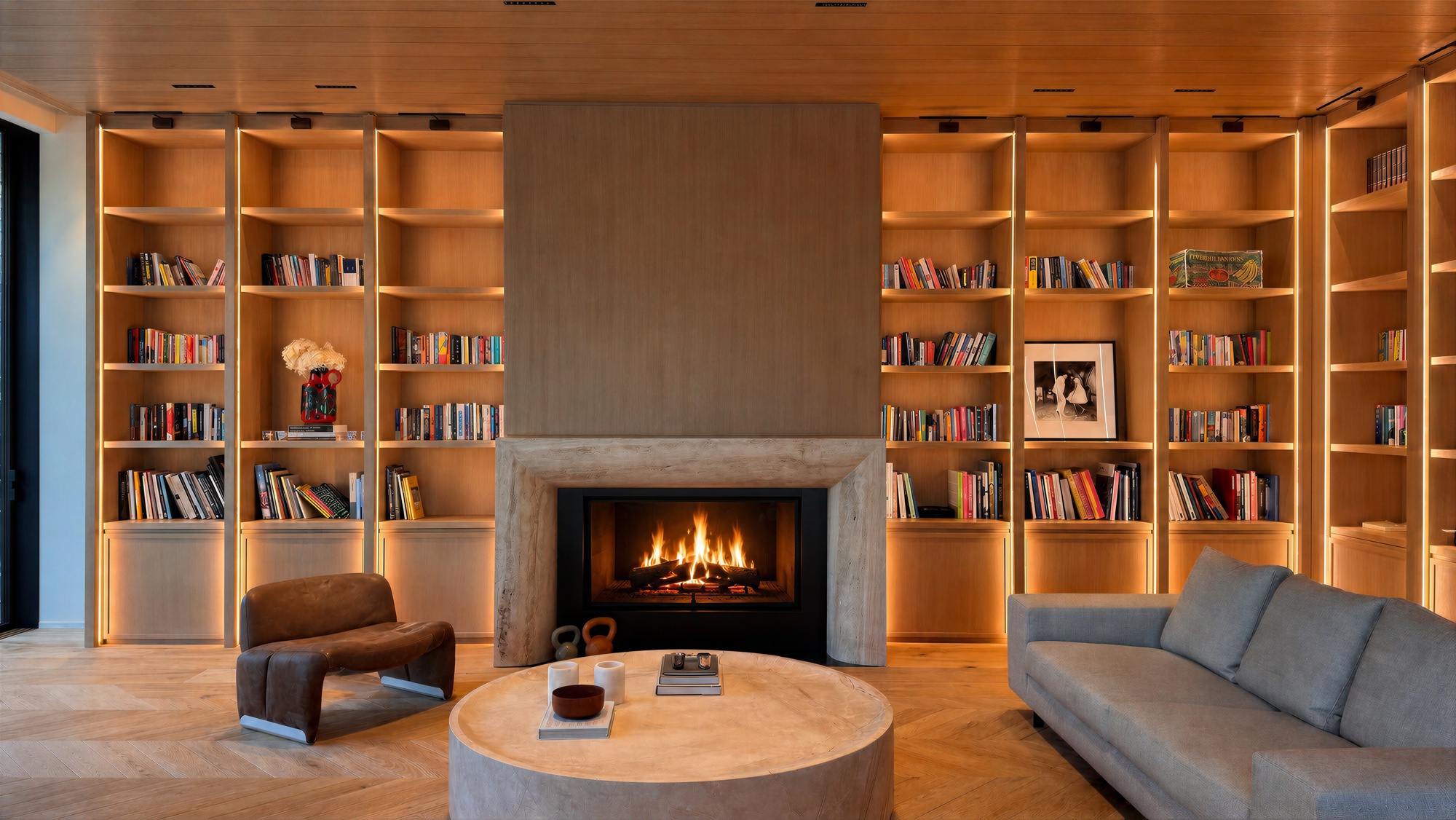 Lit fireplace in a travertine surround, flanked by built-in oak bookshelves, a round stone table and a grey sofa