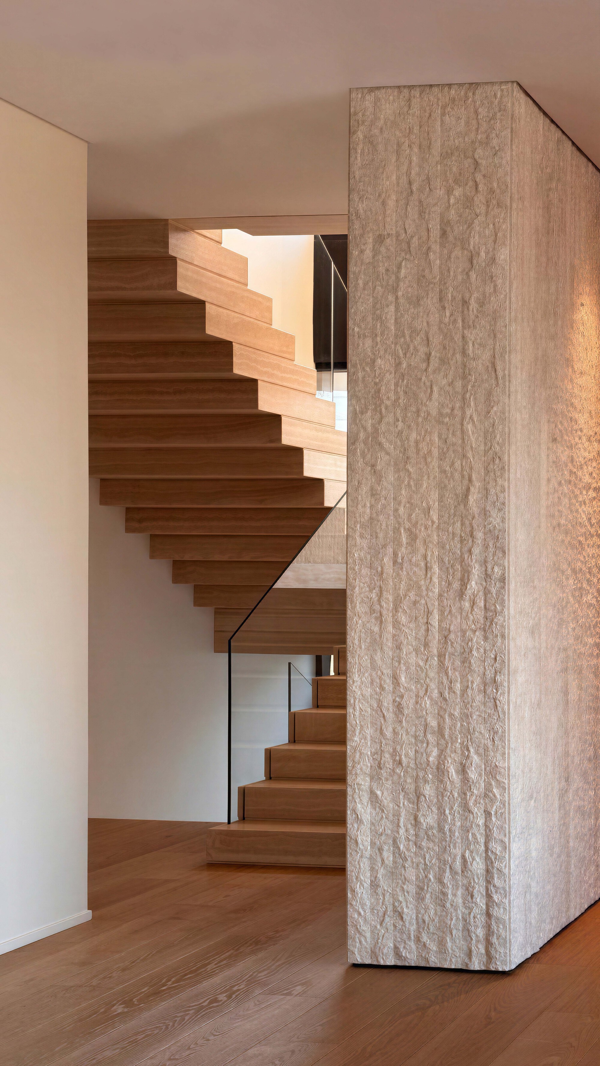 Oak staircase with glass balustrade rising beside a freestanding bush-hammered stone wall, warm light on the herringbone floor