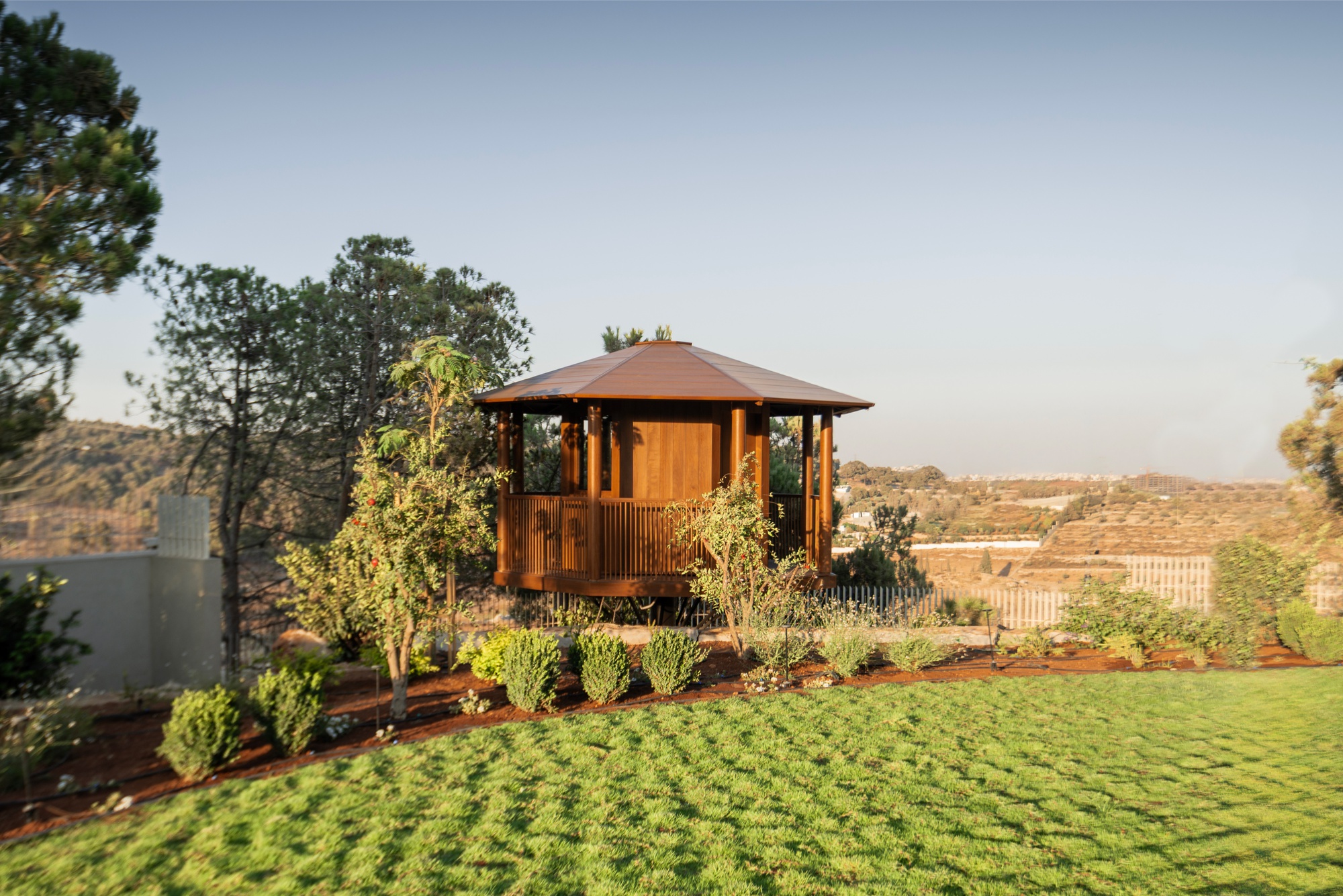 Timber gazebo with a pyramid roof on a garden hillside, with a trimmed lawn and shrubs in low evening sun