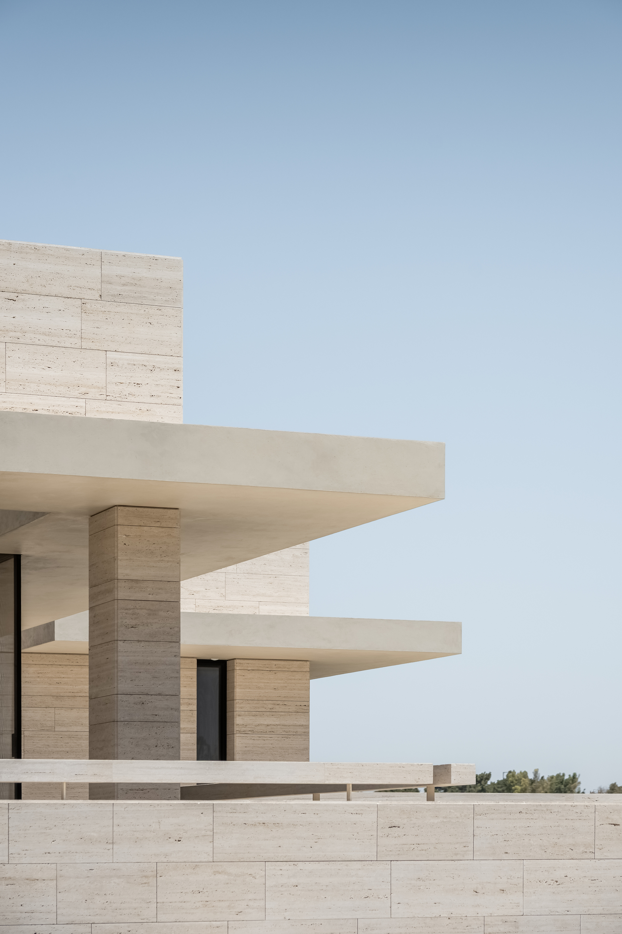 Corner of stacked travertine volumes with thin cantilevered concrete slabs and a square stone column against a clear pale sky
