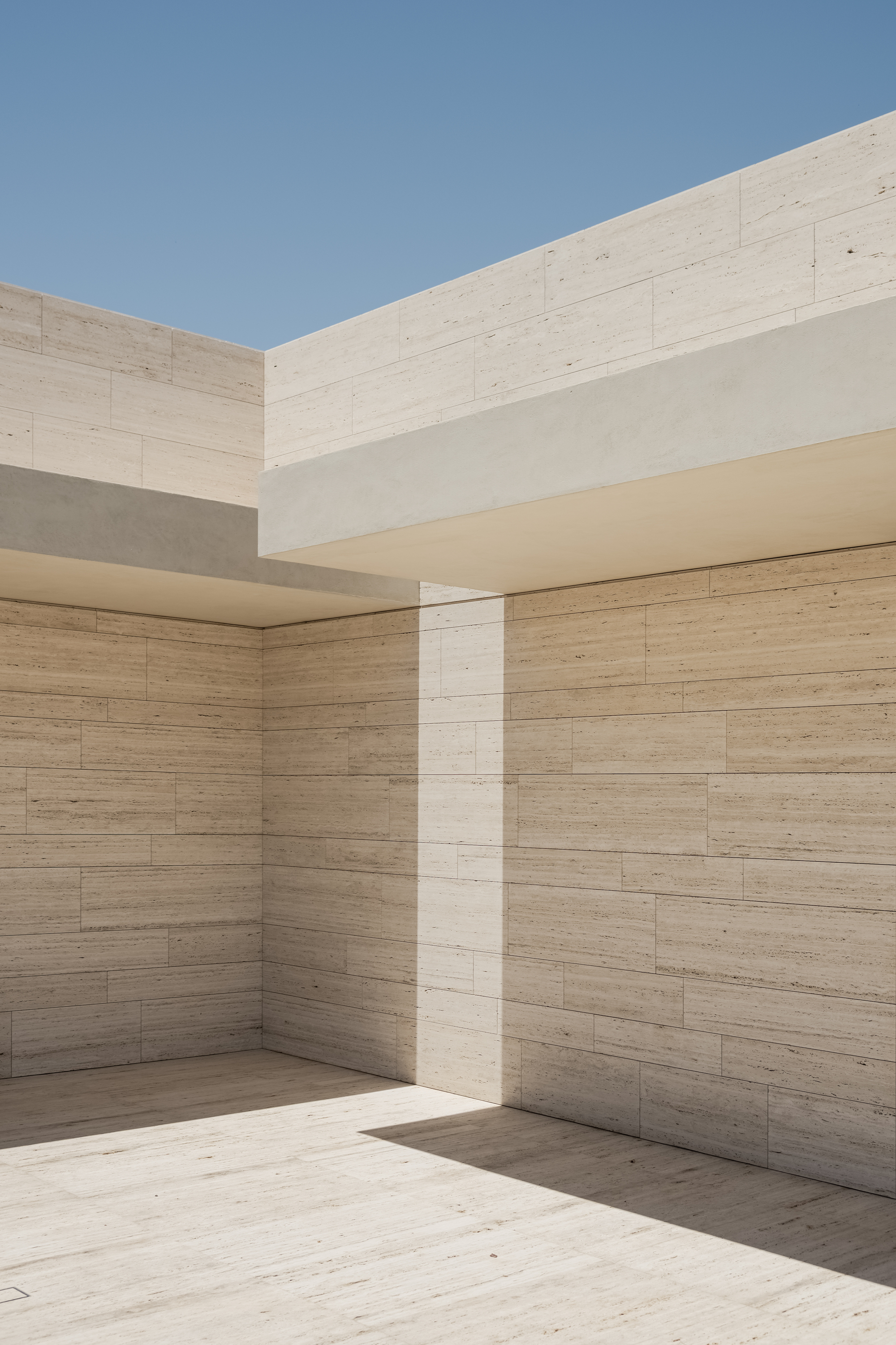 Travertine-clad courtyard corner where a cantilevered slab casts a sharp diagonal shadow across the wall and floor under clear blue sky