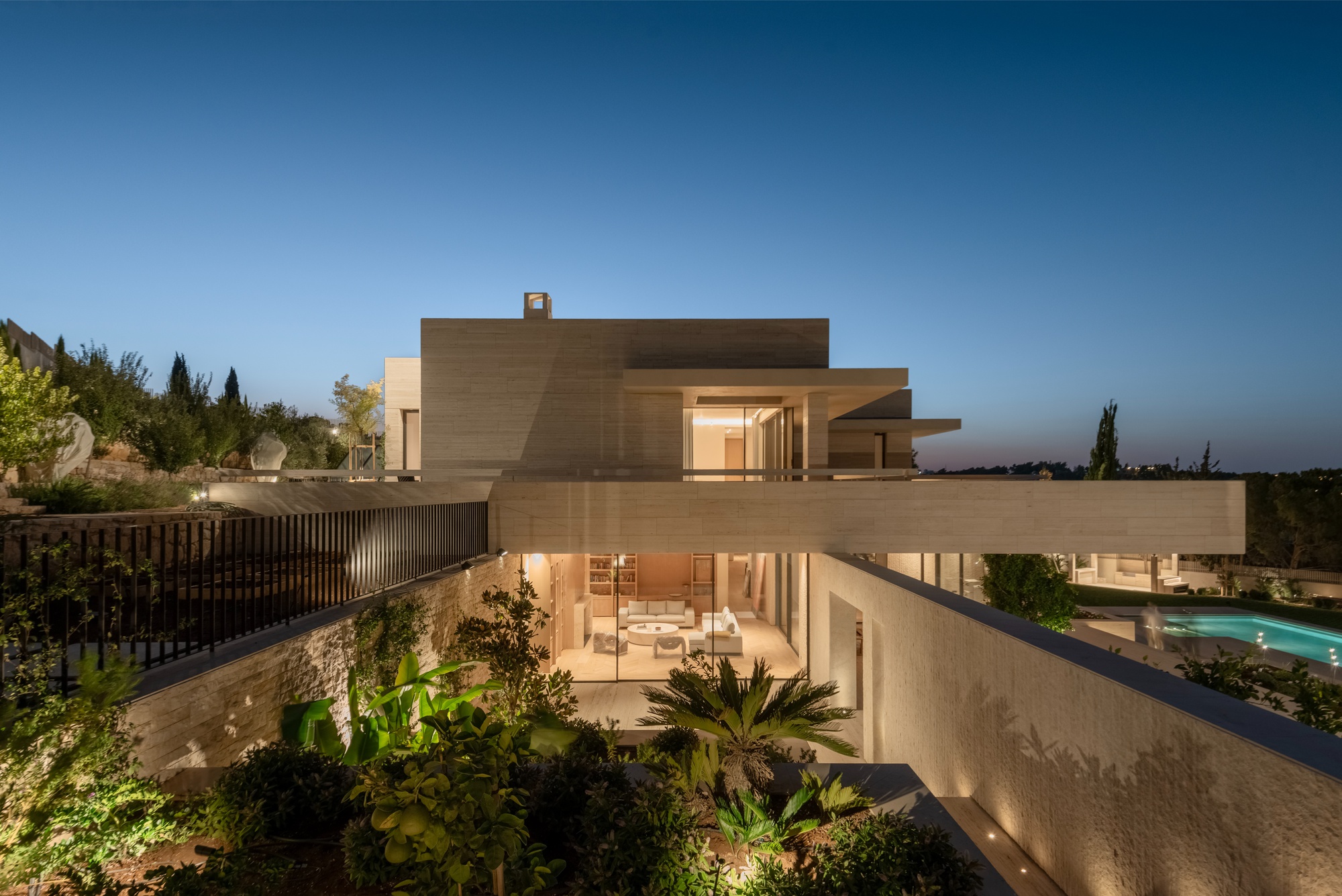 Dusk view across a planted sunken garden toward the lit glass living room, stacked stone volumes and a pool at right