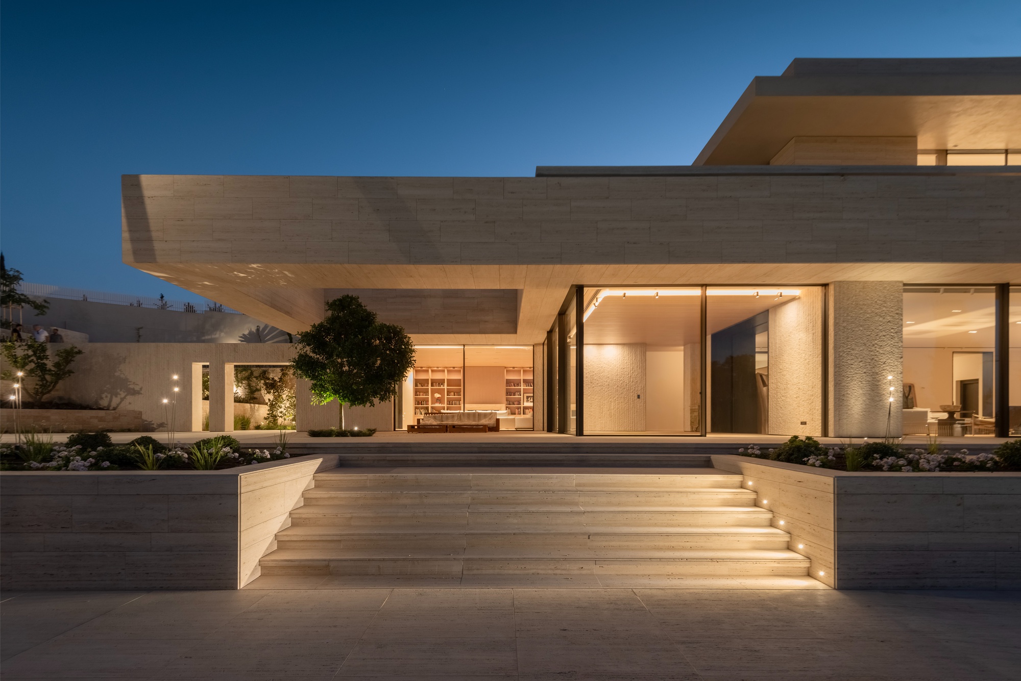 Entrance steps with lit risers between stone planters, leading to a deep cantilevered canopy and glazed living area at dusk