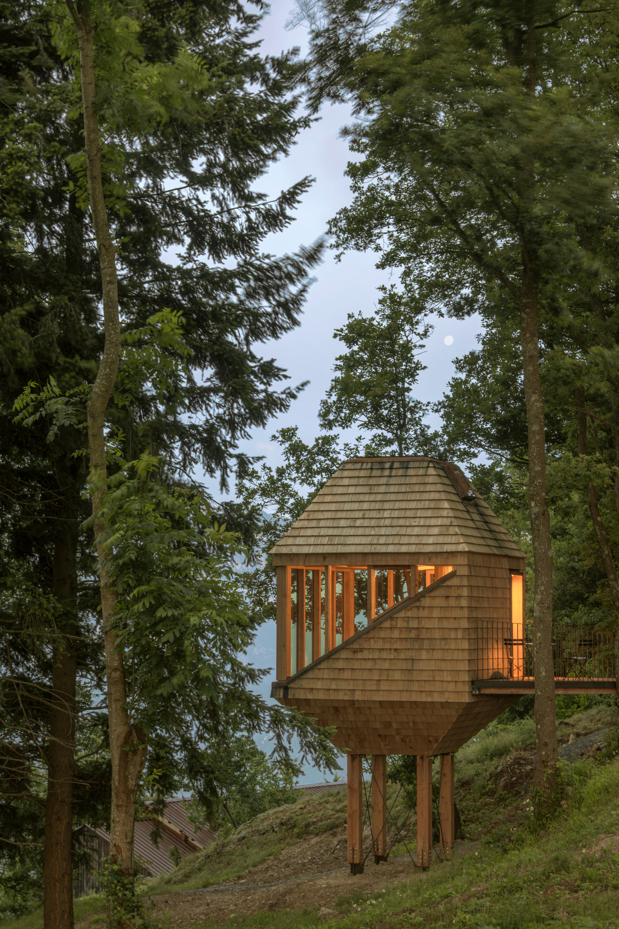 Shingled cabin raised on slender timber legs on a slope, windows glowing amber at dusk with a pale moon above