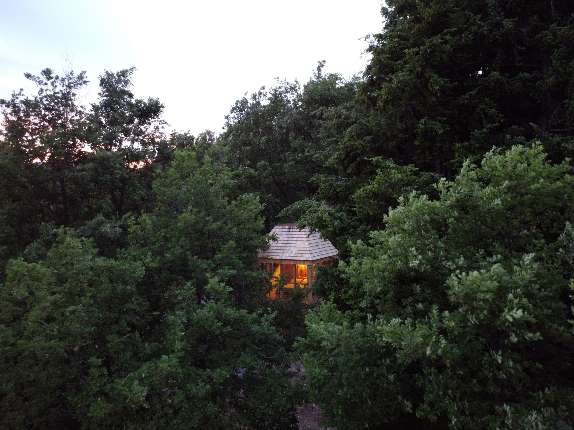 Drone view of the shingle-roofed cabin with lit windows, tucked among dense treetops at twilight