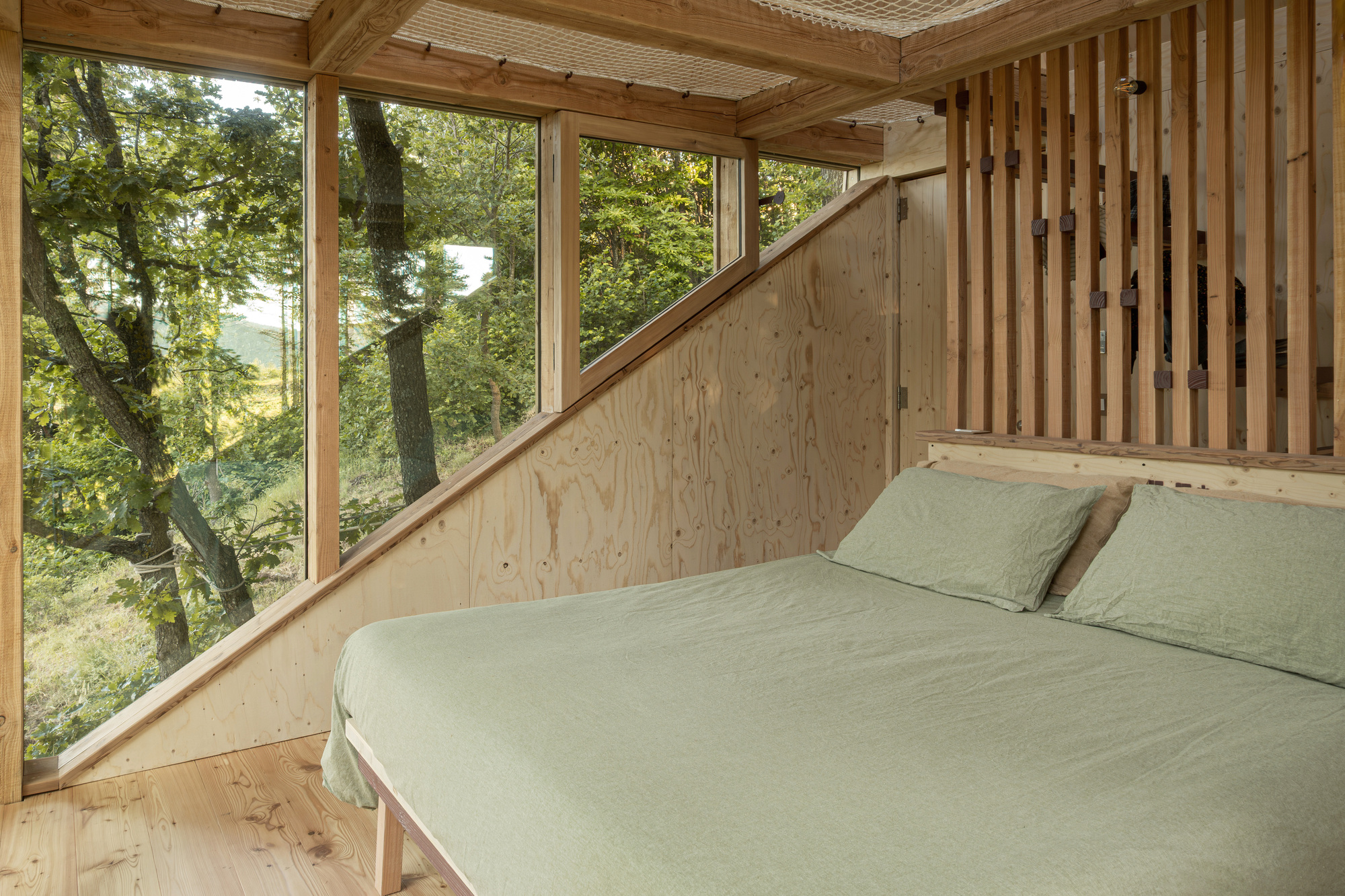 Bedroom interior with sage linen bed, plywood sloped wall, slatted timber screen and tall windows onto the trees