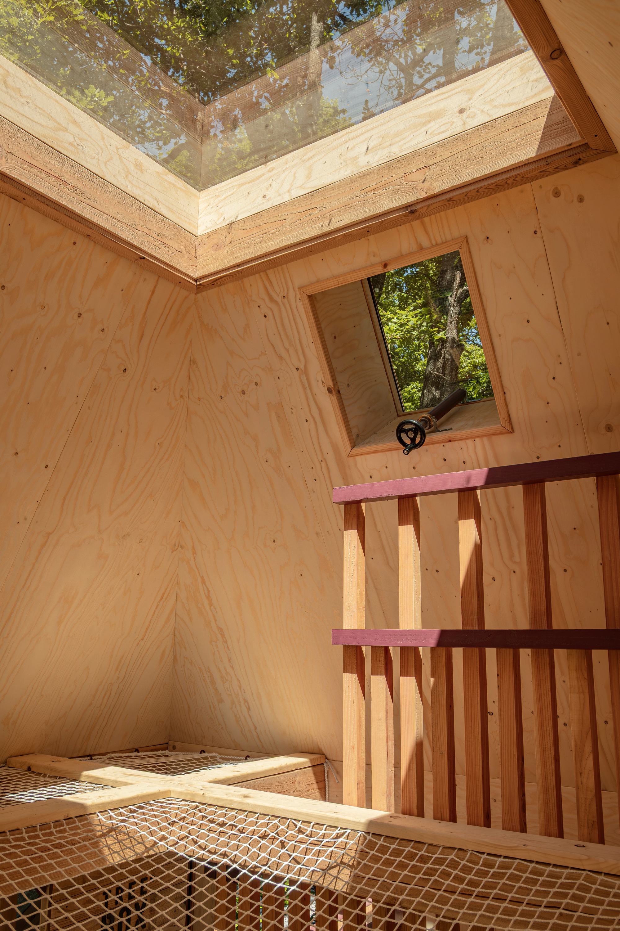 Plywood-lined corner with a roof skylight, a small opening window and a red-topped timber baluster beside a rope net floor