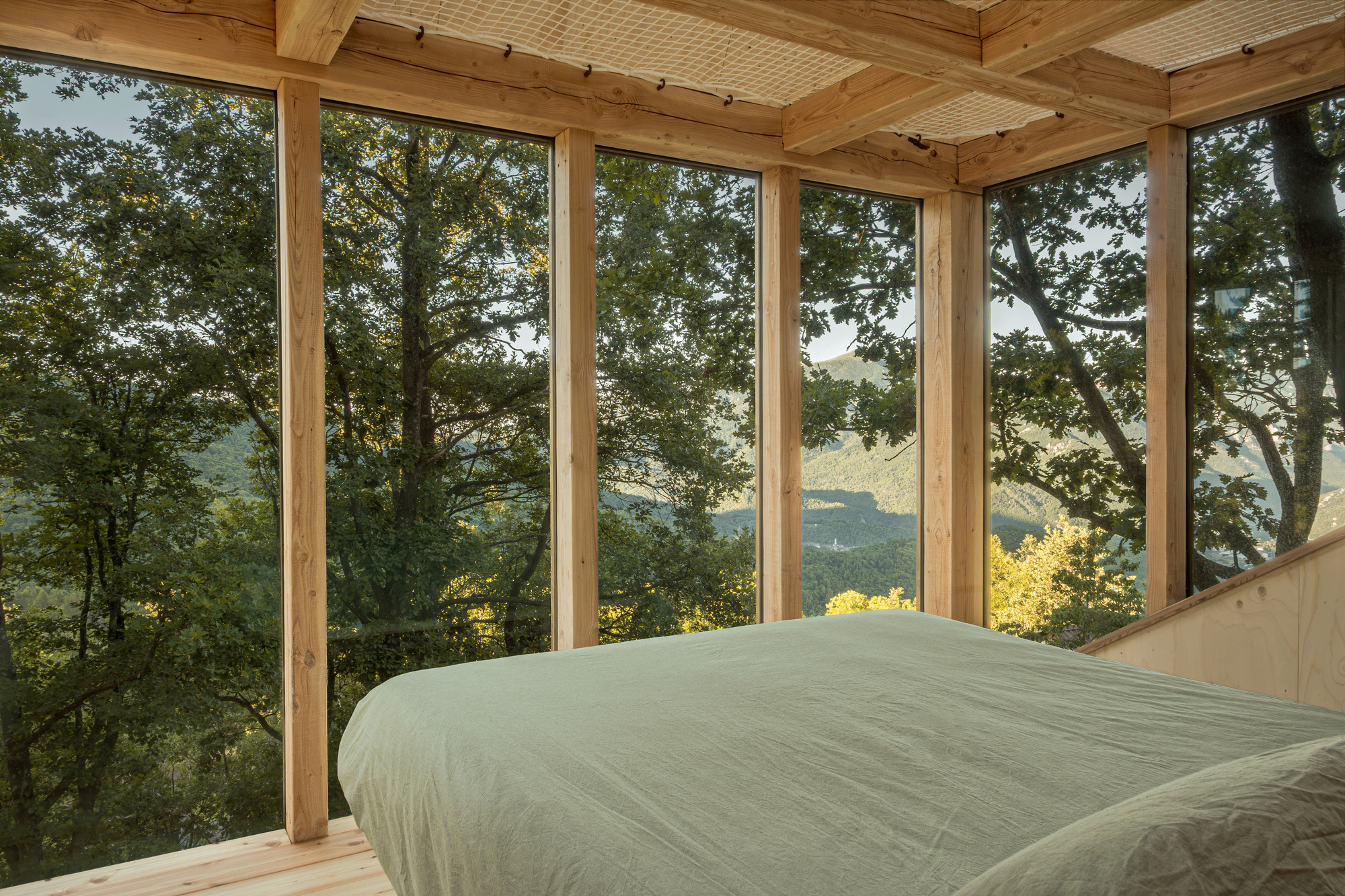 Bed beside floor-to-ceiling timber-framed glazing looking into oak canopy and distant hills in late afternoon light