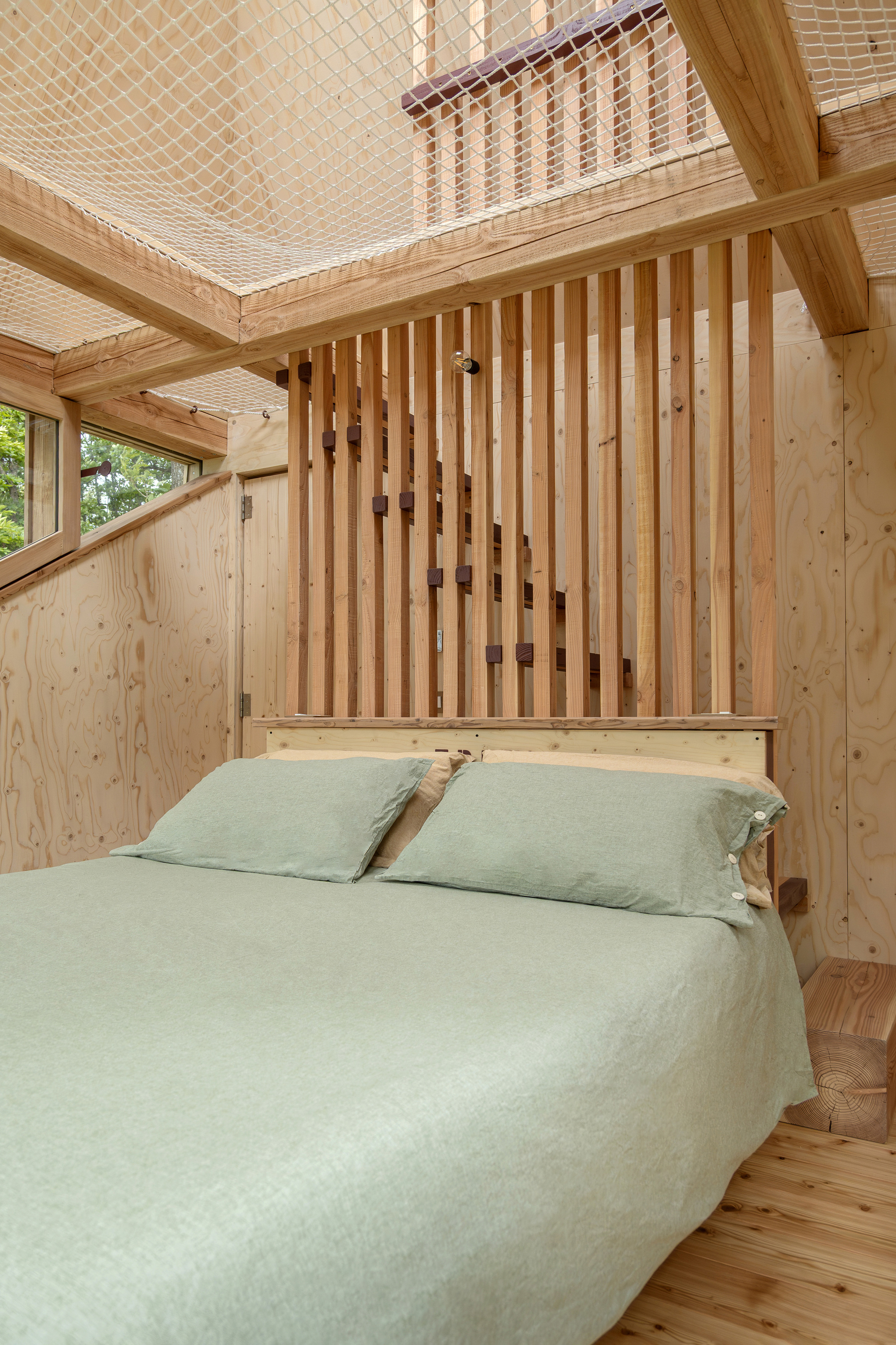 Double bed with green linen against a vertical timber slat screen, under a rope net ceiling and exposed beams